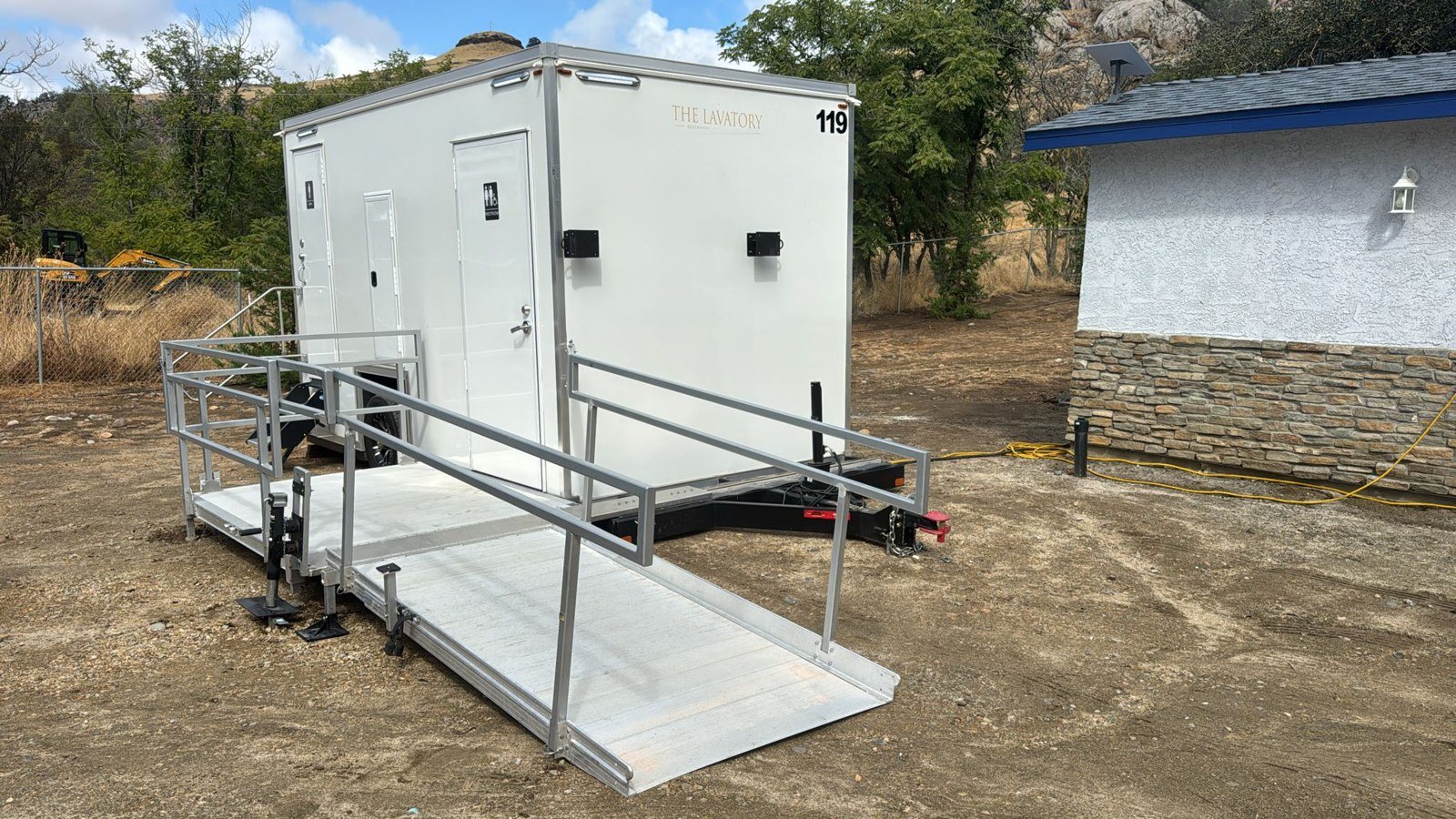 A Lavatory accessible restroom trailer with a long aluminum ramp and rails on a dirt job site, with an excavator behind the fence