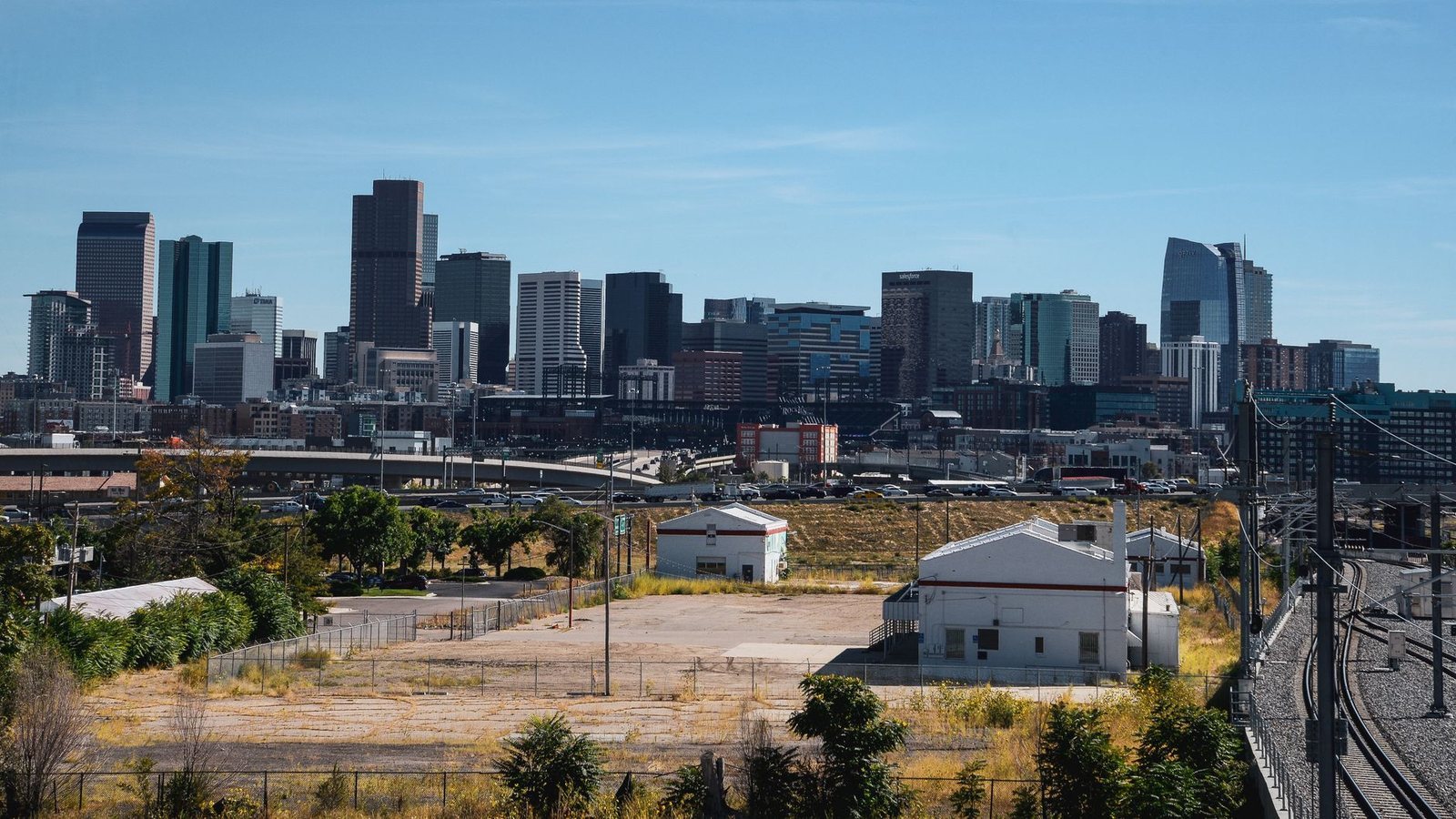 Downtown Denver skyline on a clear day, seen across an open lot and rail yard north of the freeway