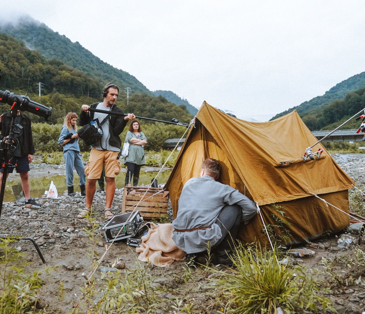 A film crew with a boom microphone and lights shooting beside a canvas tent on a rocky riverbank in a mountain valley