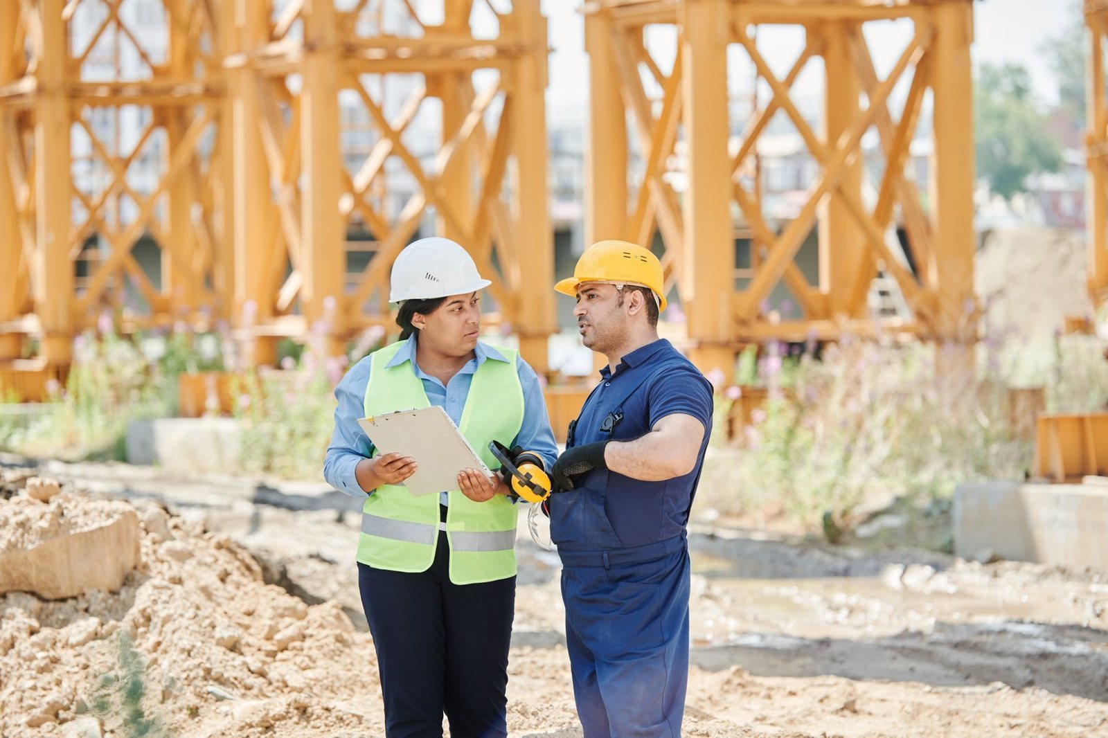 Two construction workers in hard hats and safety vests talking over a clipboard on a dirt job site with steel framing behind them