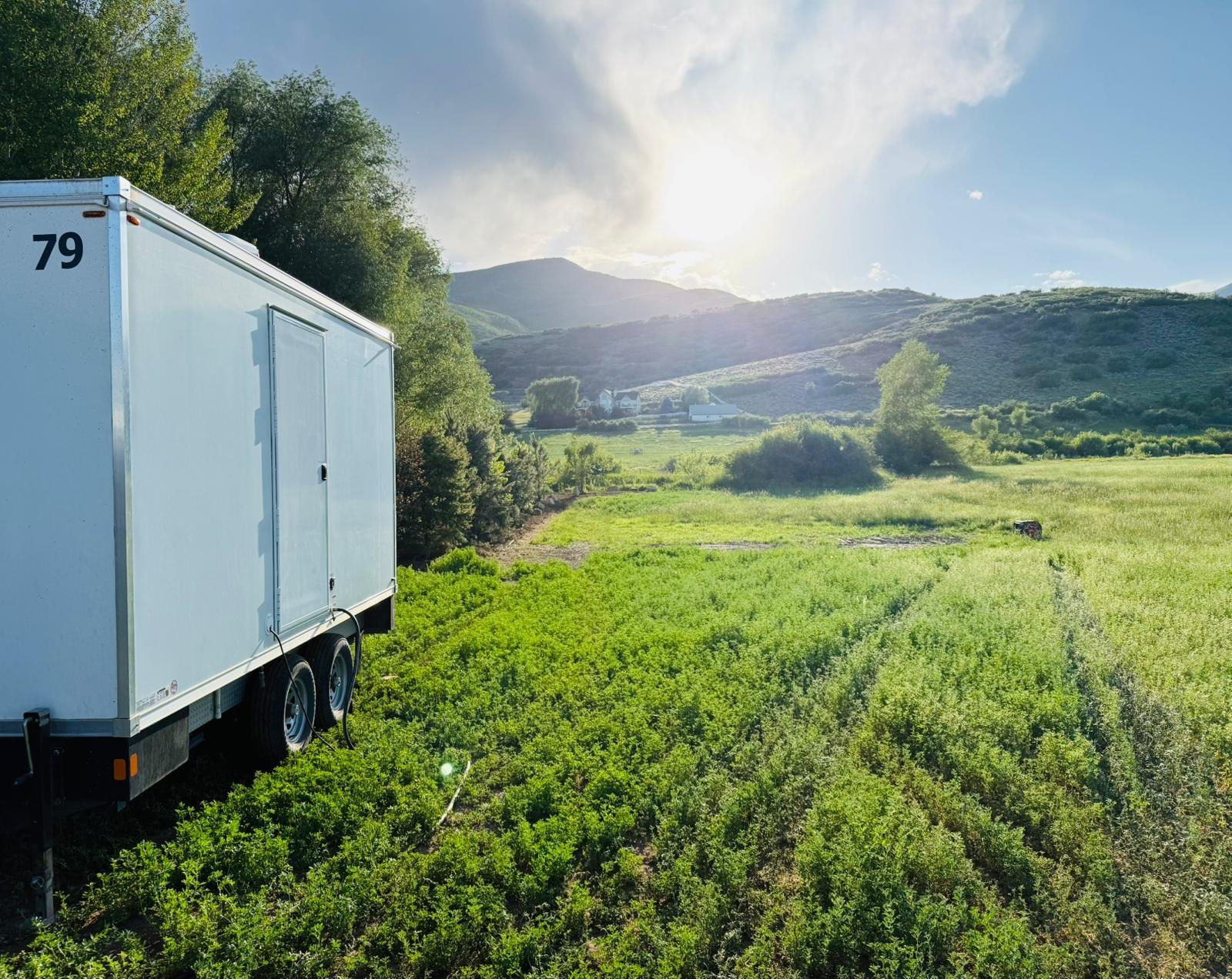 A Lavatory restroom trailer parked at the edge of a green mountain meadow, with foothills, a farmhouse and late sun behind it