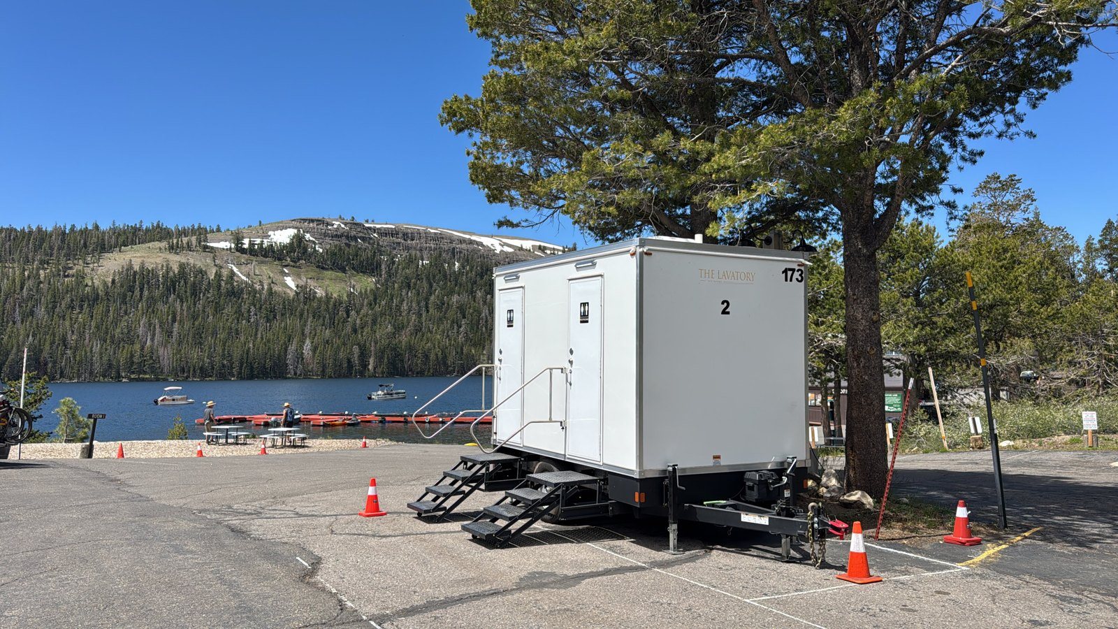 A Lavatory 2-Station restroom trailer under a pine tree in a paved lot beside a mountain lake, with two people near picnic tables on the shore and boats on the water
