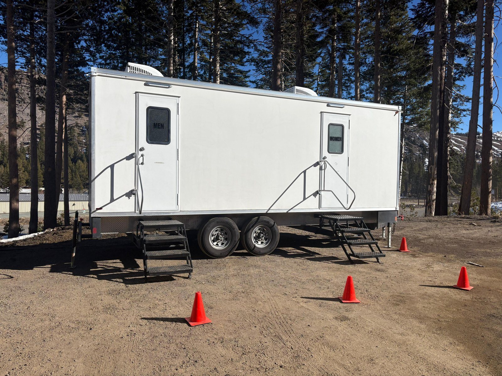 A Lavatory 2-Station restroom trailer with men's and women's doors and steel stairs, set on a dirt lot among tall pines with traffic cones around it