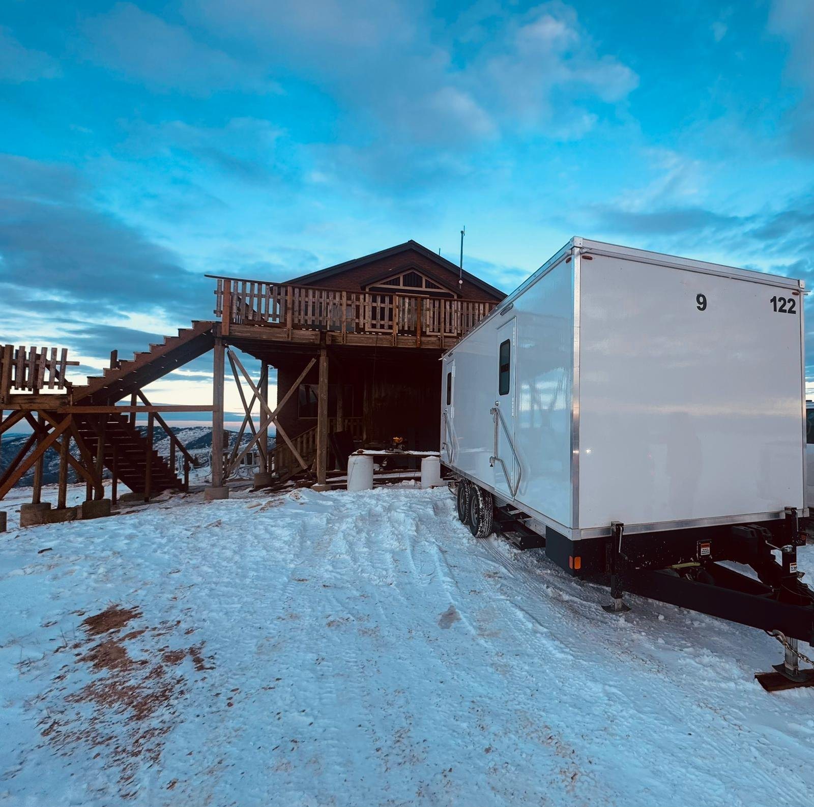 A Lavatory 9-Station restroom trailer parked on packed snow beside a timber lodge with an outdoor staircase at a high mountain site