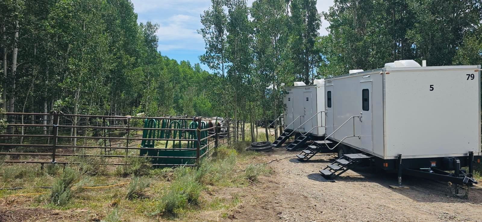 Two Lavatory 5-Station restroom trailers lined up on a gravel drive in an aspen grove next to a horse corral
