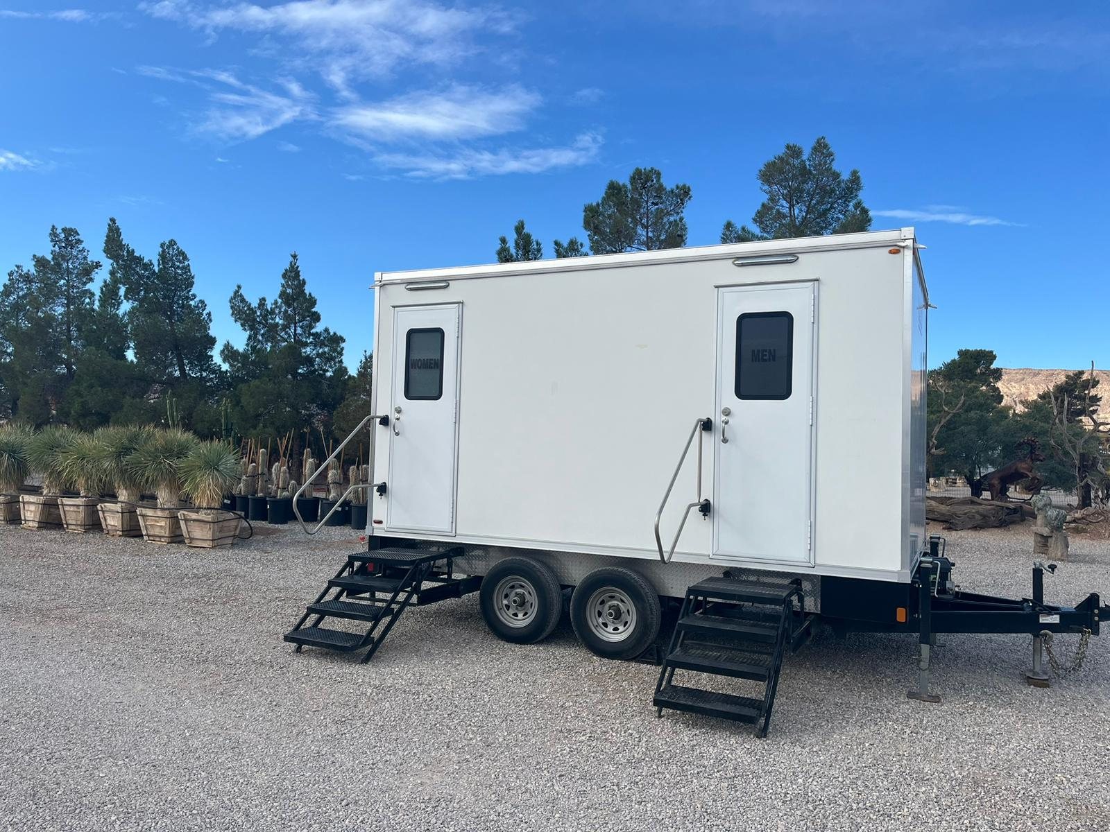 A Lavatory 2-Station bathroom trailer on a gravel lot with pine trees and a clear blue sky behind it