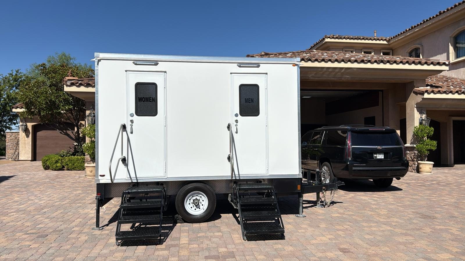 A Lavatory 2-Station restroom trailer parked on the paver drive of a large home, with a SUV beside it