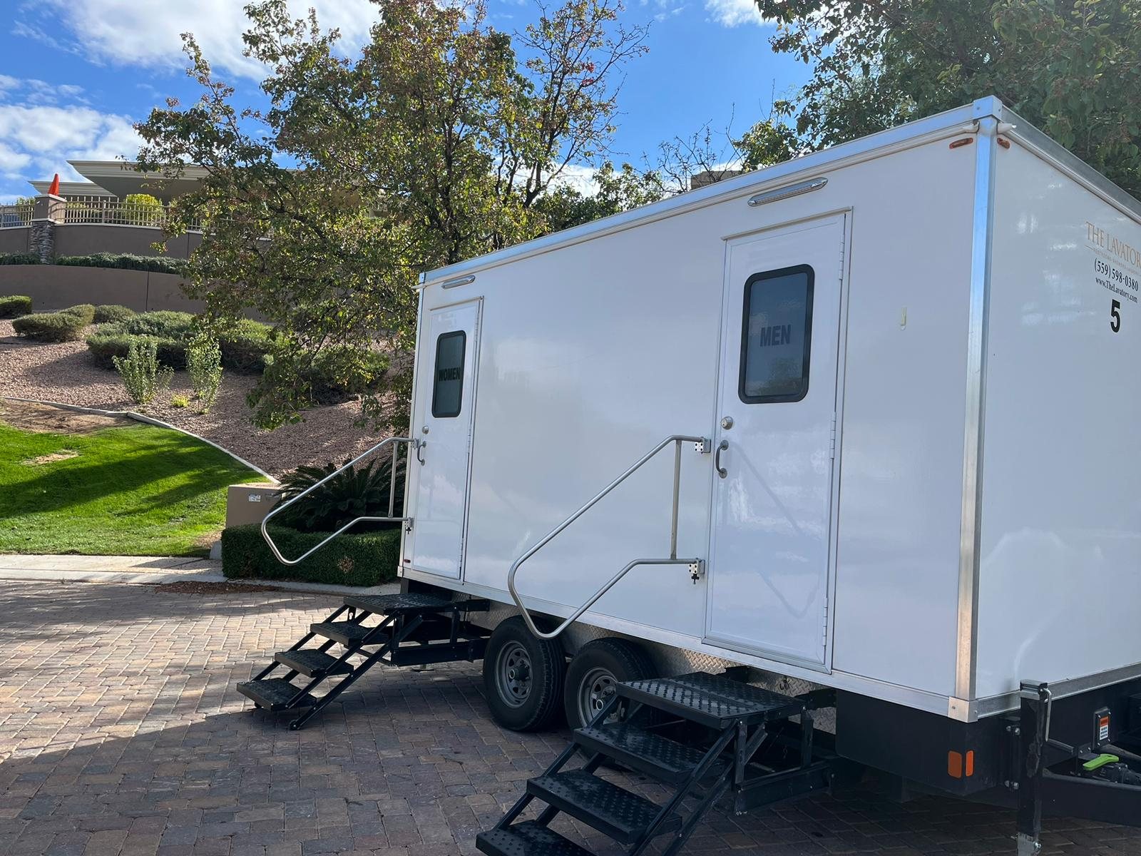 A Lavatory 5-Station restroom trailer on a paver driveway beside a green lawn and shade trees, stairs set at each door