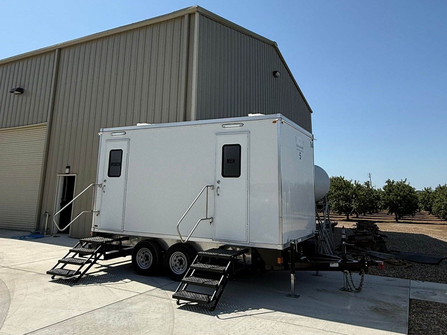A Lavatory restroom trailer with women's and men's doors set on a concrete pad beside a metal farm building, with rows of orchard trees behind it