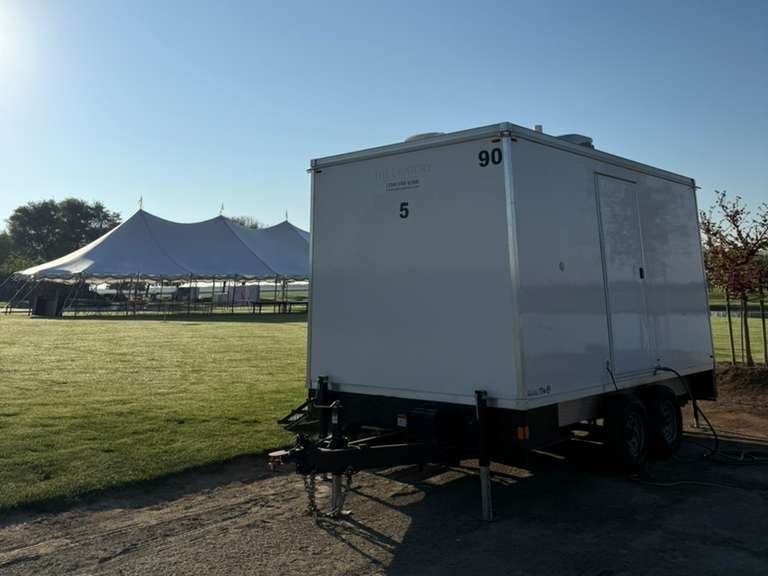 A Lavatory 5-Station restroom trailer parked on a gravel edge beside a green lawn, with a large white event tent set up behind it in the early morning sun