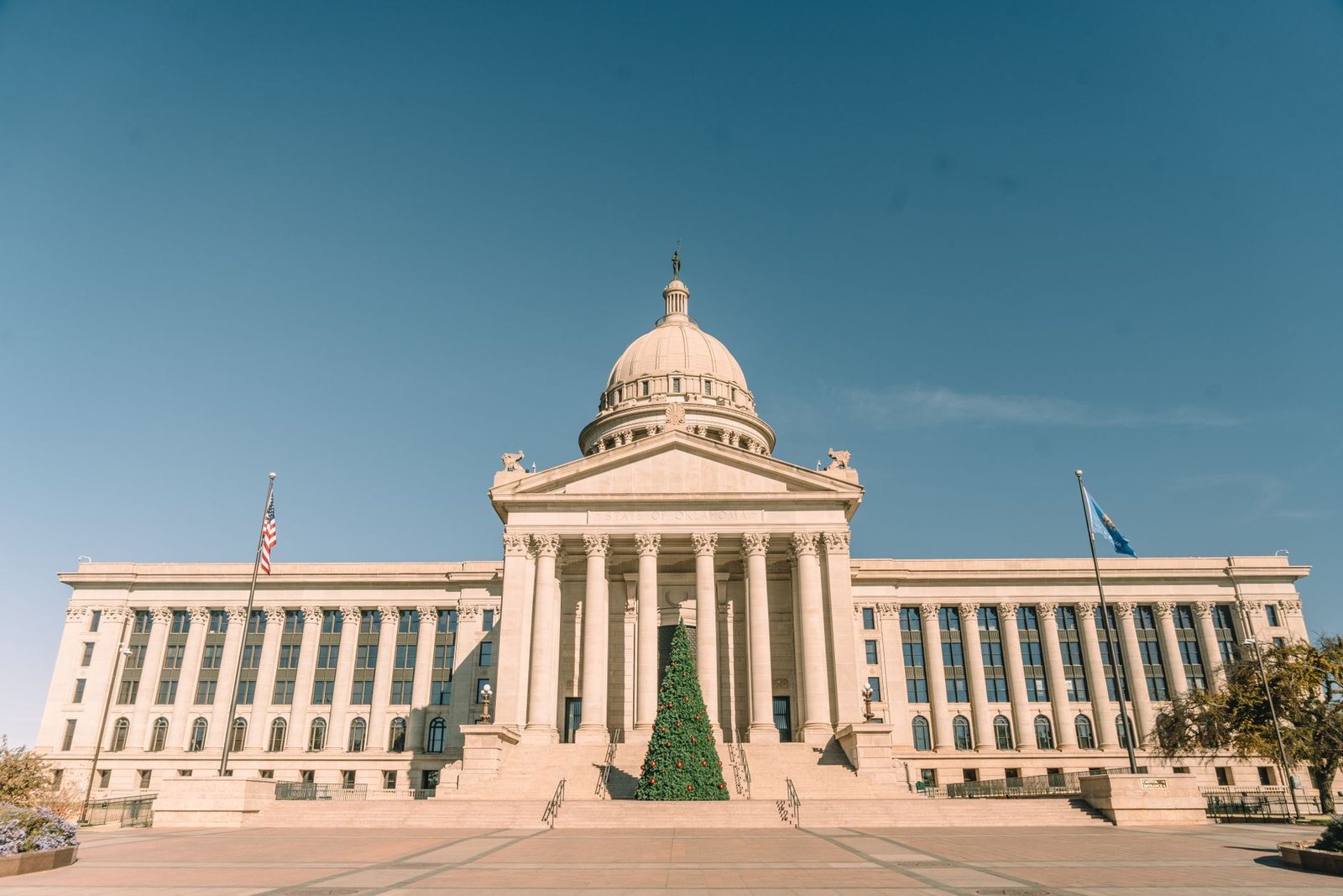 The Oklahoma State Capitol in Oklahoma City under a clear blue sky, with a holiday tree on the front steps