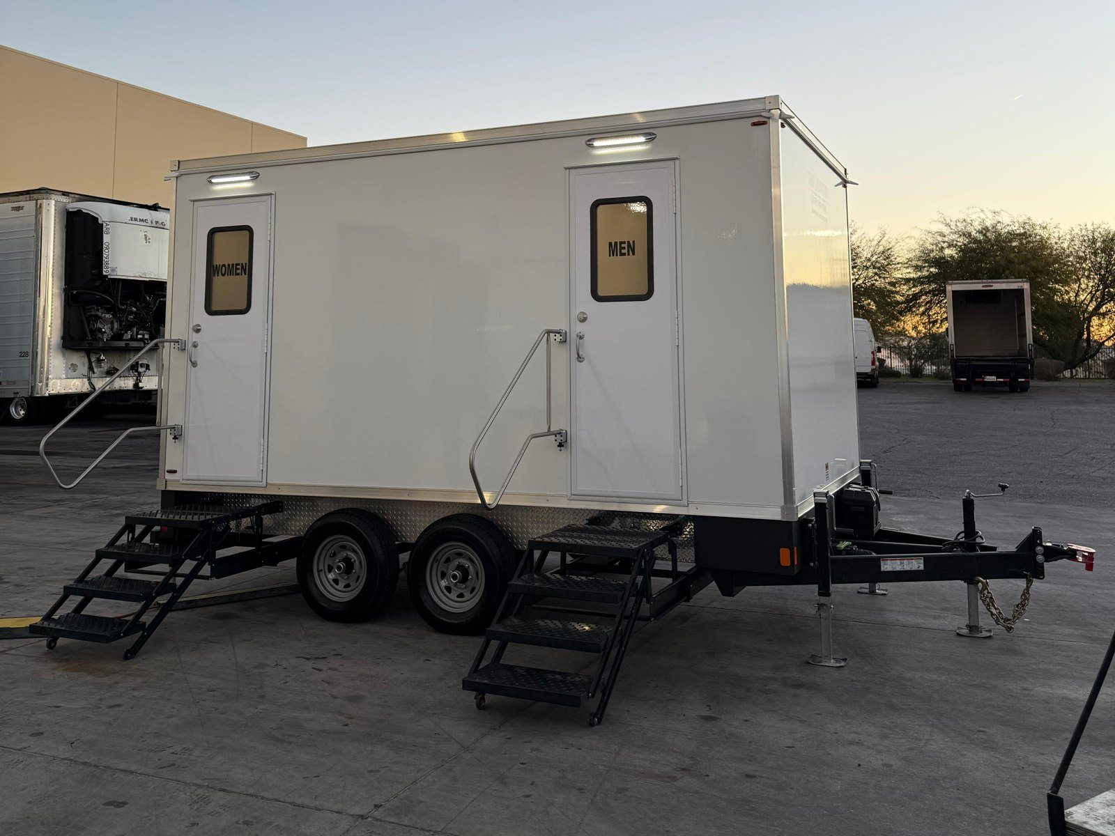 A Lavatory restroom trailer with WOMEN and MEN doors and lit exterior lights, set in a paved industrial yard at dawn beside parked box trucks