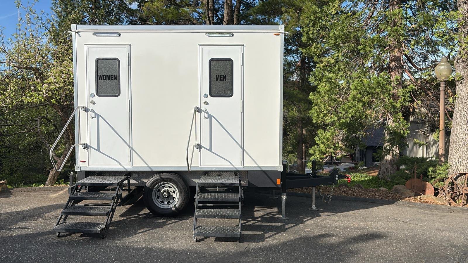 A Lavatory 2-Station restroom trailer with WOMEN and MEN doors and steel stairs, set on a paved drive under tall pines and a vintage lamp post
