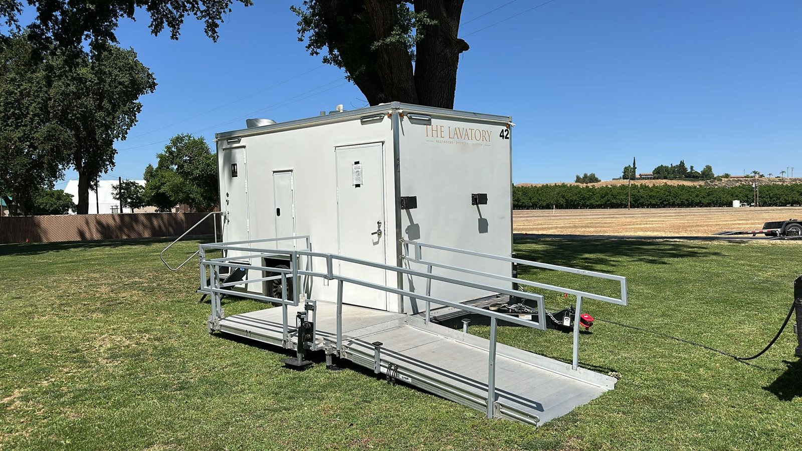 A Lavatory ADA restroom trailer with a long aluminum ramp and handrails set up on an open lawn under a large shade tree