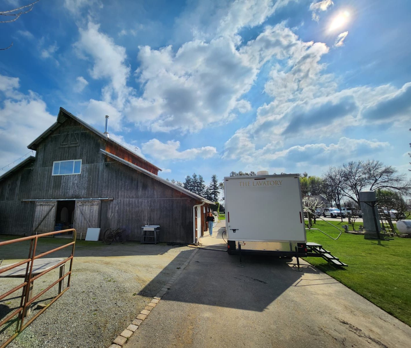 A Lavatory restroom trailer parked on the drive beside a weathered wooden barn wedding venue under a big cloudy sky