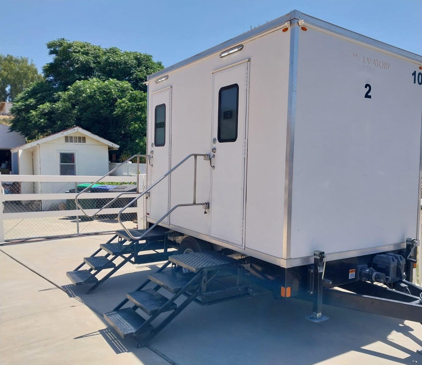 A Lavatory 2-Station restroom trailer with women's and men's doors set on a concrete home driveway next to a white fence and garden shed