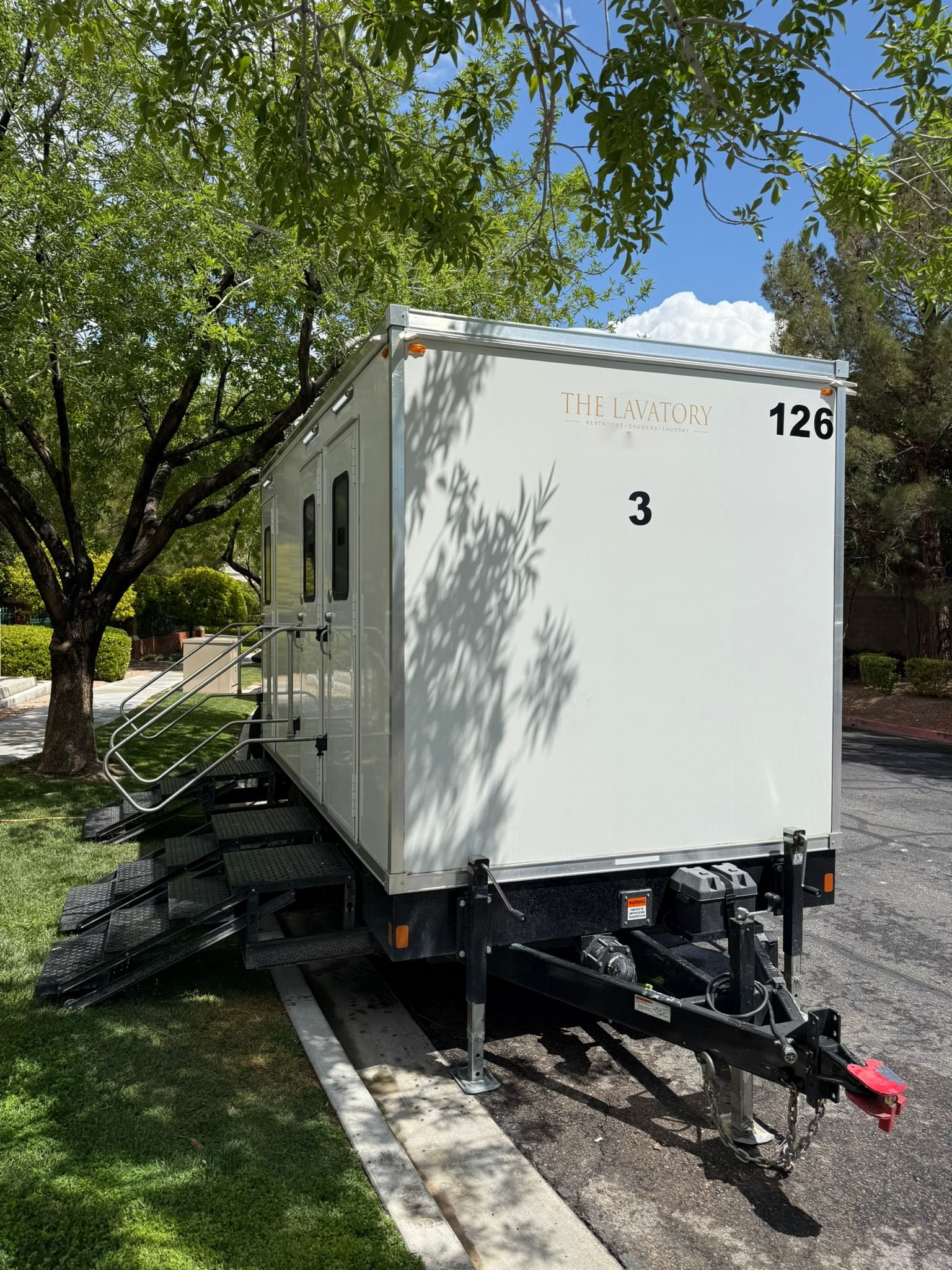 A Lavatory 3-Station restroom trailer with steel stairs parked along a shaded park lawn beside a paved lot