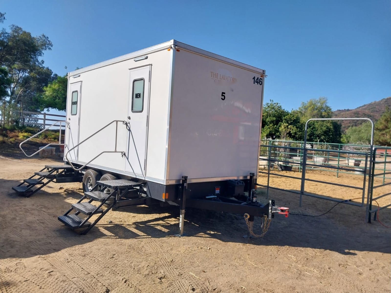 A Lavatory 5-Station restroom trailer with separate women's and men's doors and steel stairs, parked on a dirt lot beside metal horse pens at a ranch