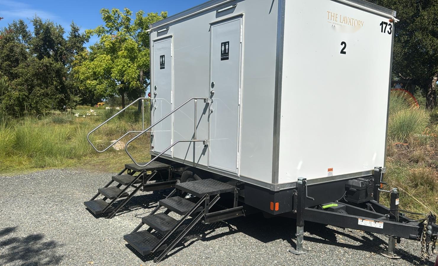 A Lavatory 2-Station restroom trailer with two sets of steel stairs on a gravel pad next to tall grass and leafy trees