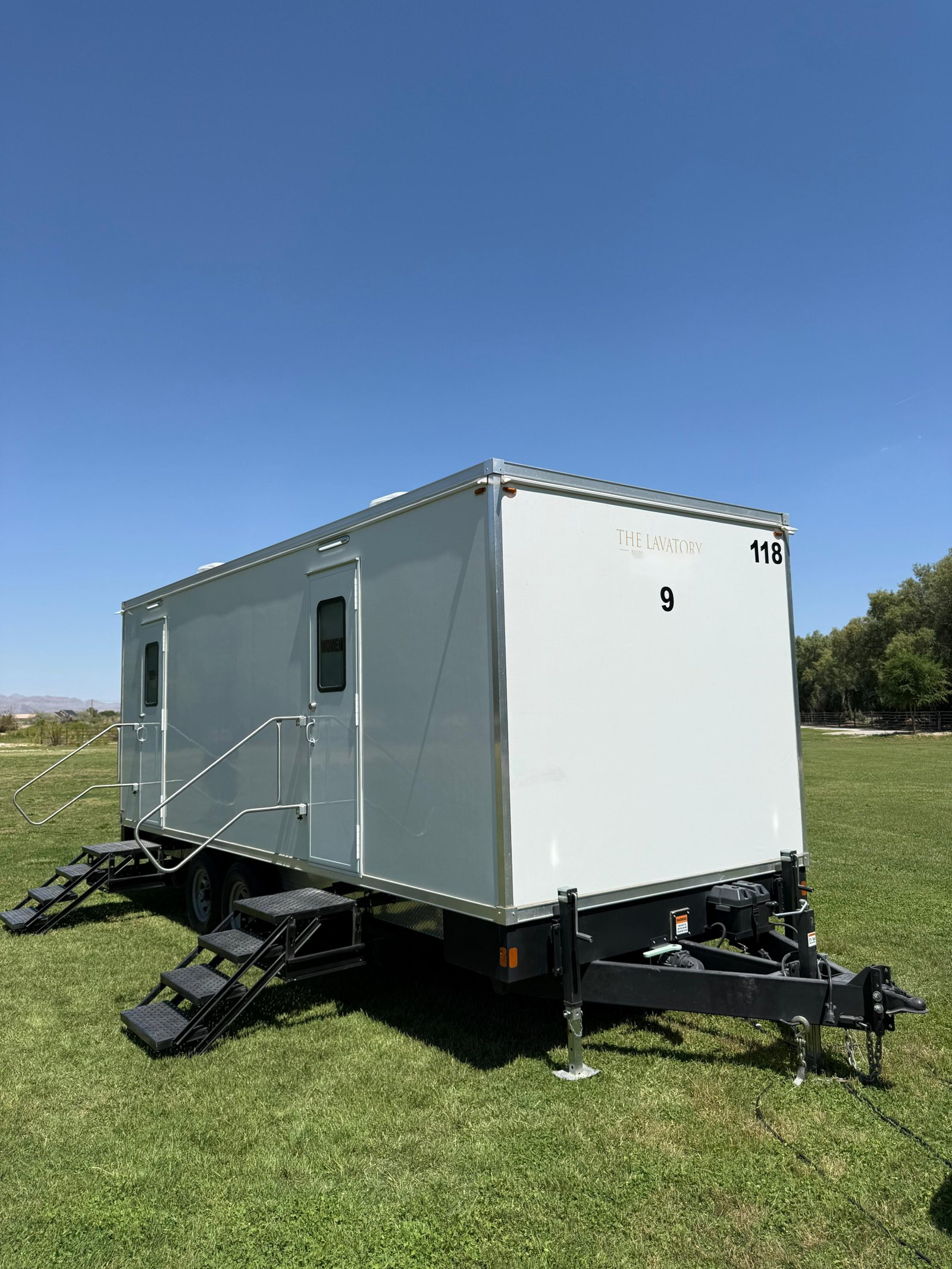 A long Lavatory 9-Station restroom trailer with two entry stairs parked on a green lawn under a clear blue sky
