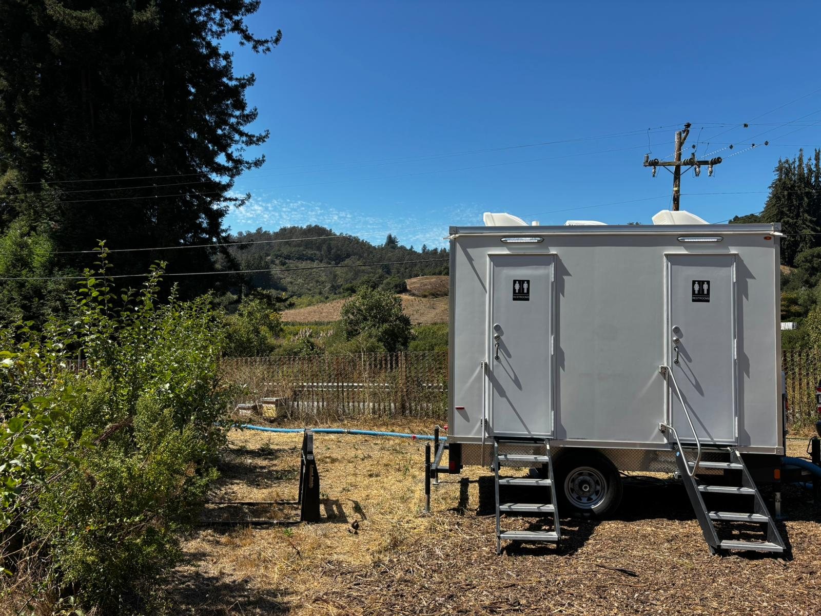 A white 2-Station restroom trailer with two restroom doors and steel stairs on a dry grass lot by a wood fence, with wooded hills and a power pole behind