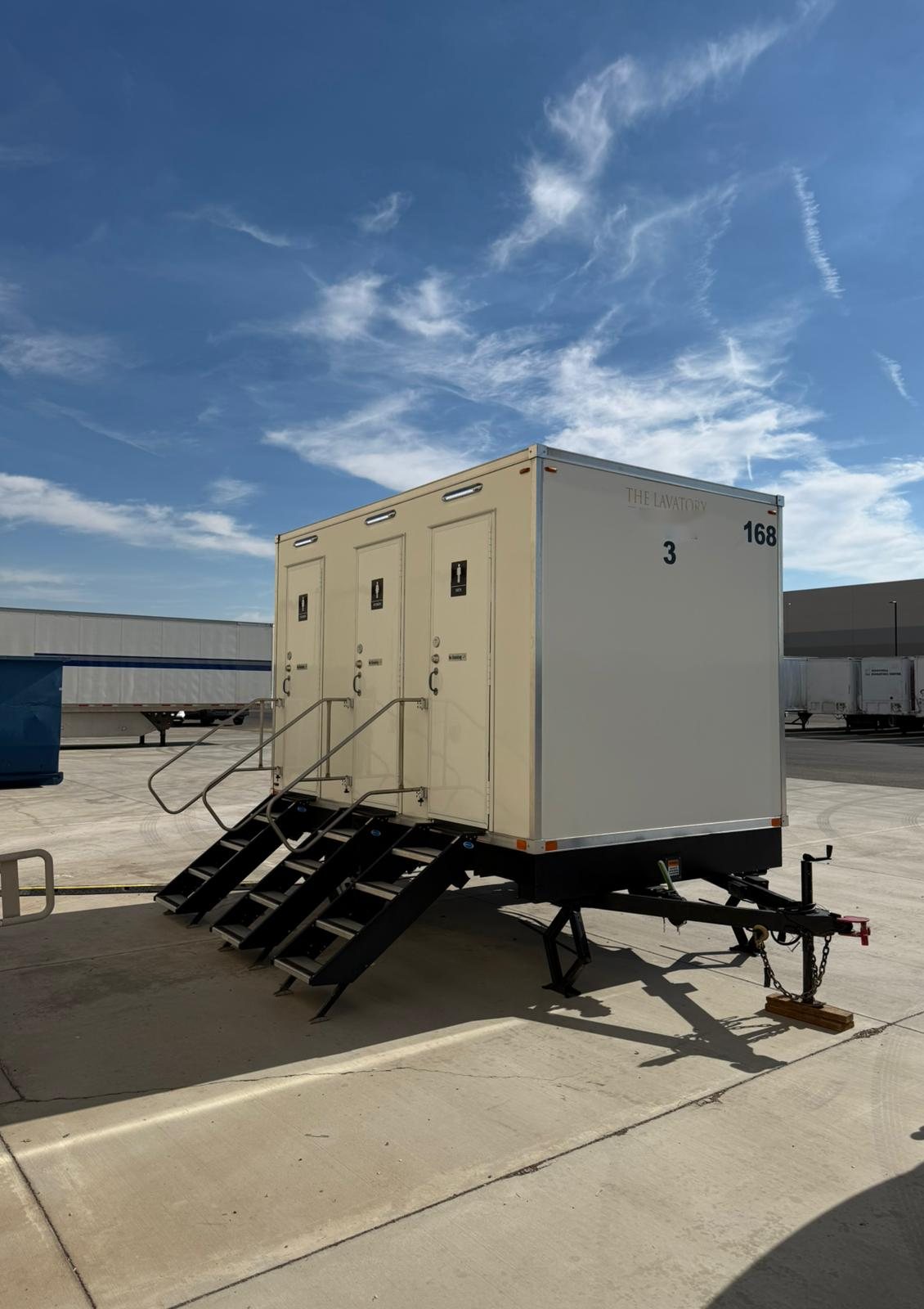 A Lavatory 3-Station restroom trailer with three doors and steel stairs set on a concrete lot beside warehouse buildings and parked semi trailers
