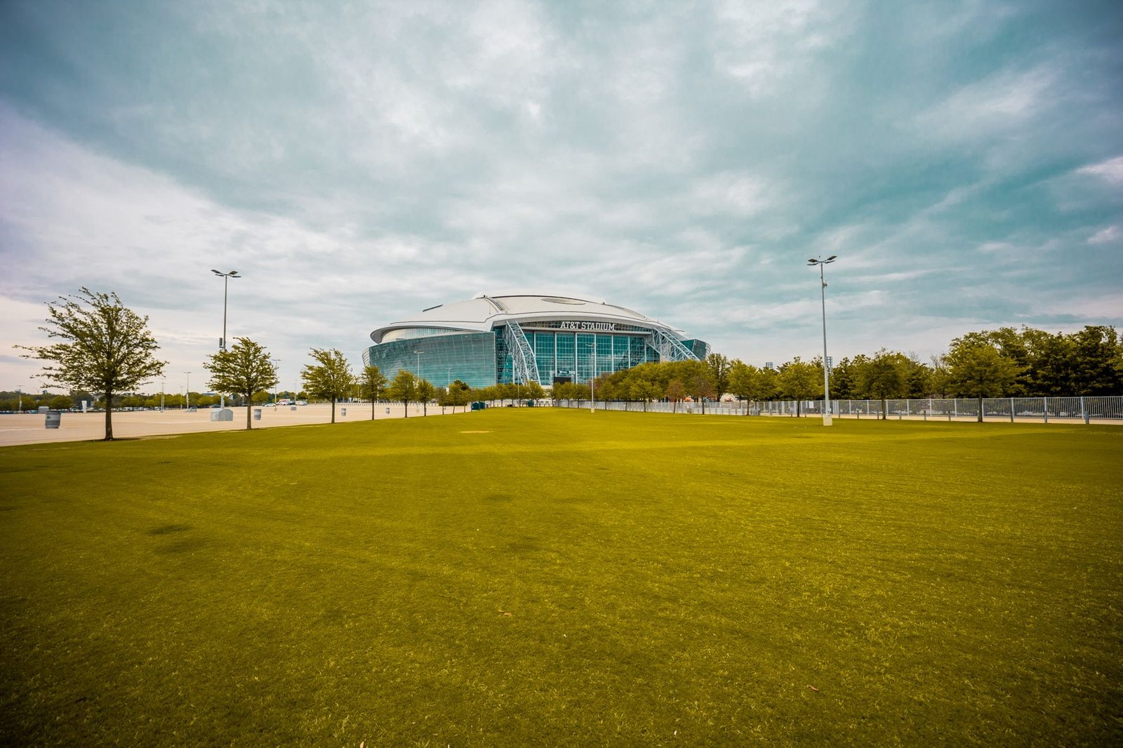 AT&T Stadium in Arlington, Texas seen across a wide grass lot lined with young trees and light poles