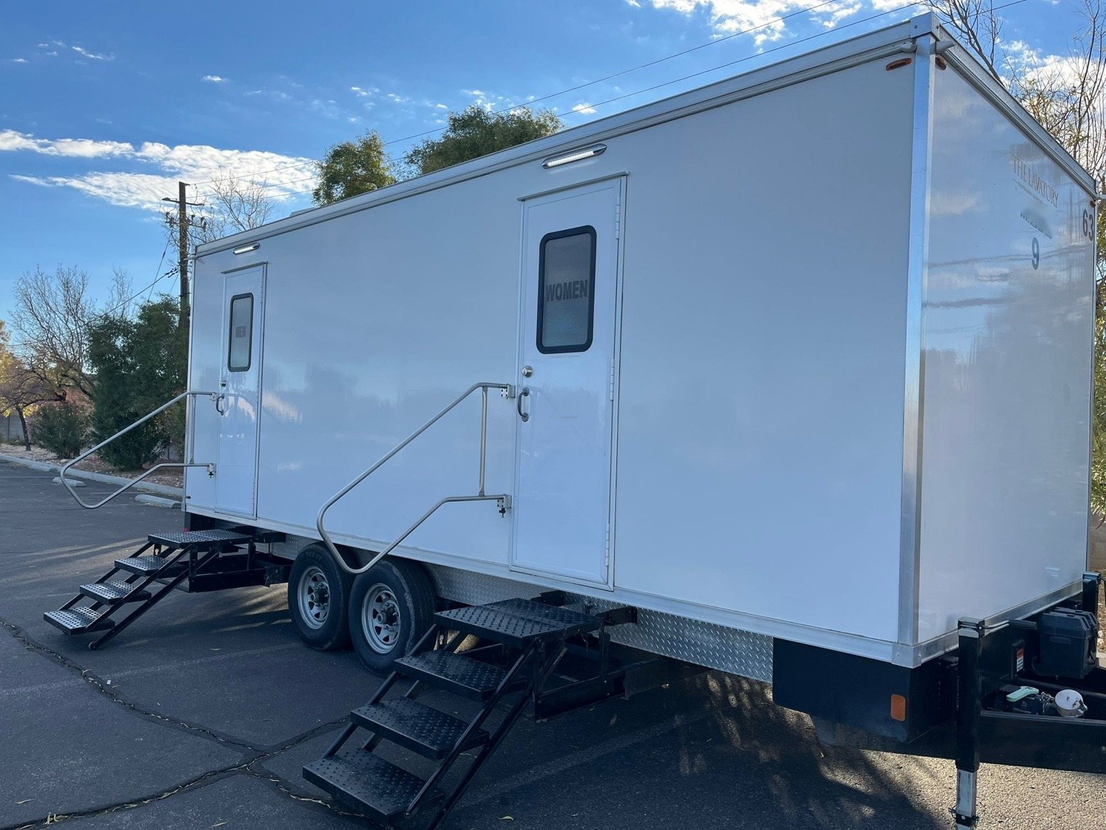 A Lavatory 9-Station restroom trailer with steel stairs and handrails parked in a paved parking lot under a blue sky