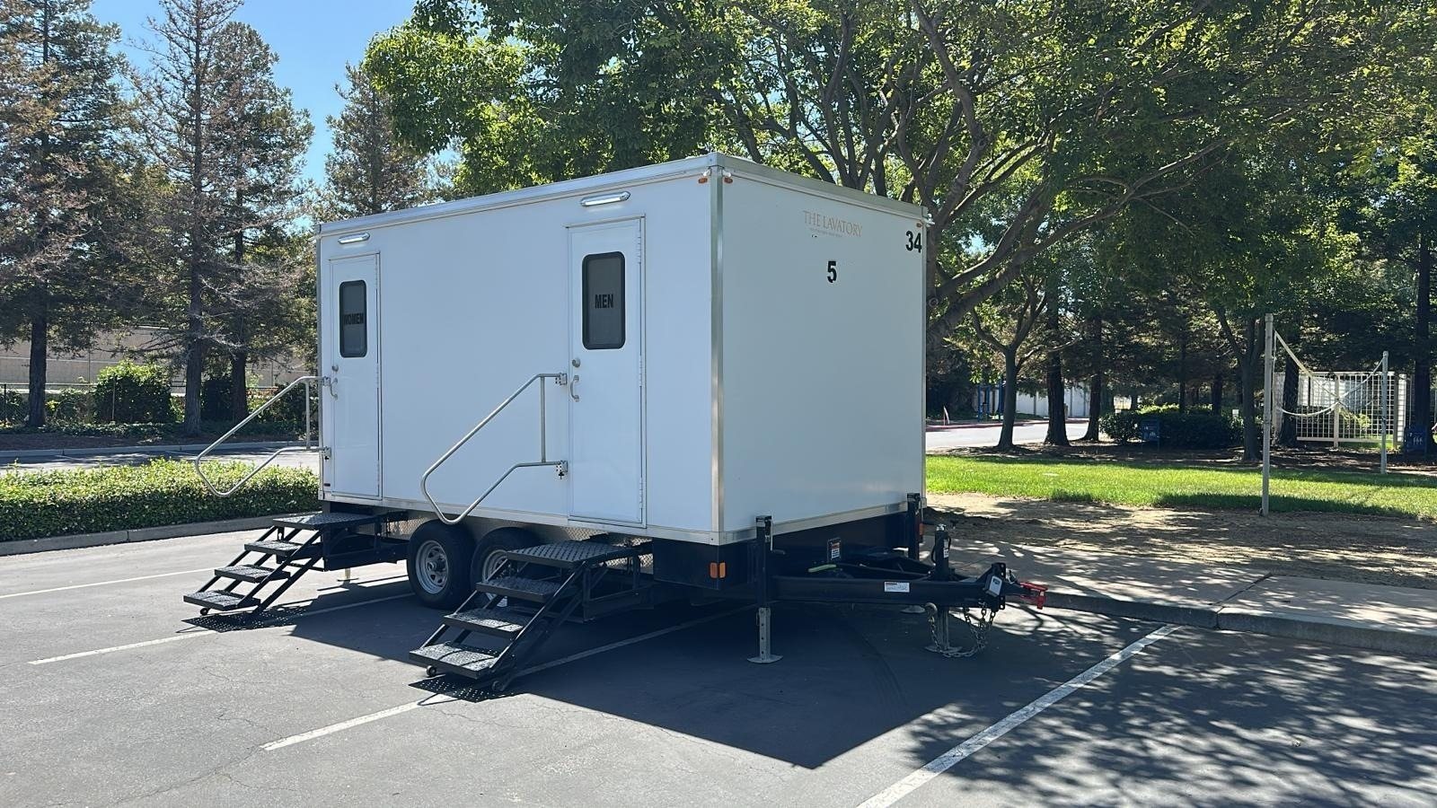 A Lavatory 5-Station restroom trailer with steel stairs parked in a shaded lot beside a grass field and tall trees