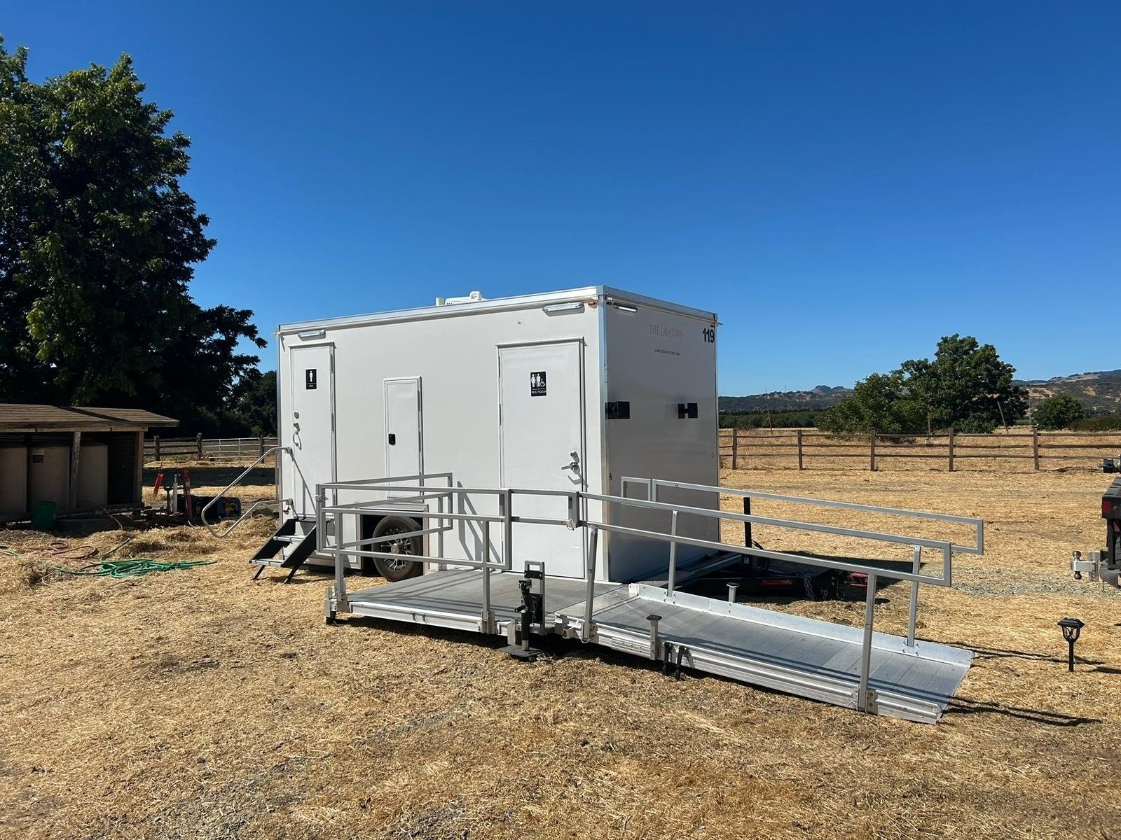 A Lavatory ADA restroom trailer with an aluminum ramp and handrails set on dry grass at a ranch with a wood rail fence and shade trees