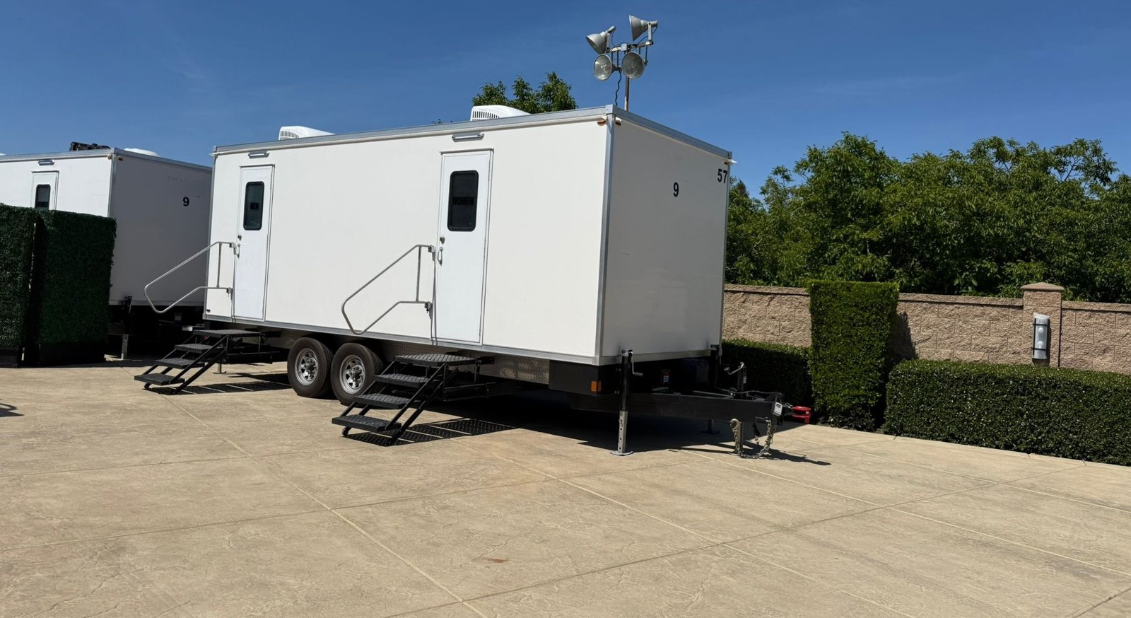 A Lavatory 9-Station restroom trailer with women's and men's doors and two steel stair sets on a paved event lot beside hedges, with a second trailer and a light tower behind it