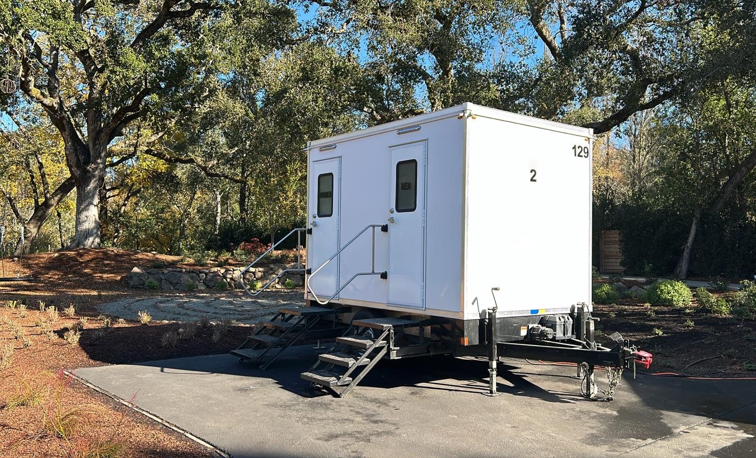 A Lavatory 2-Station restroom trailer with women's and men's doors on a paved drive under large oak trees at a garden venue