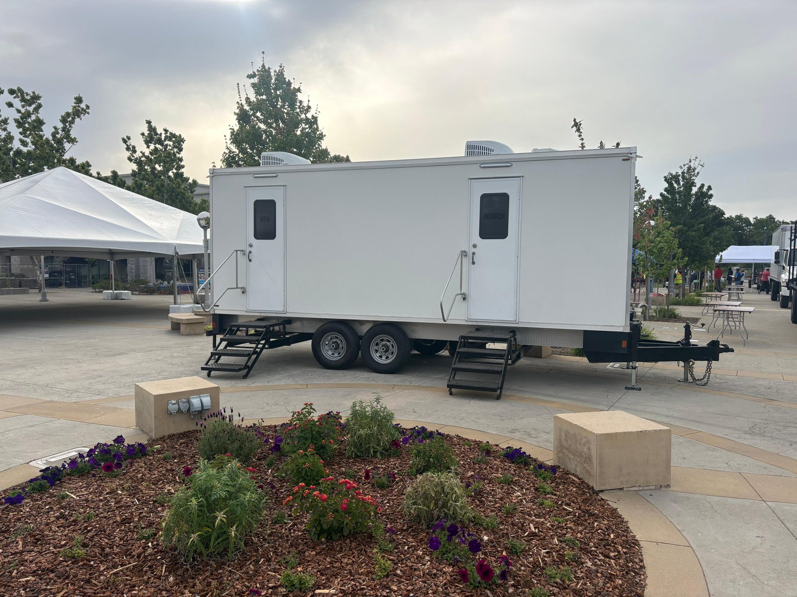 A Lavatory restroom trailer with men's and women's doors set on a paved plaza beside a large white event tent, flower beds and vendor tables