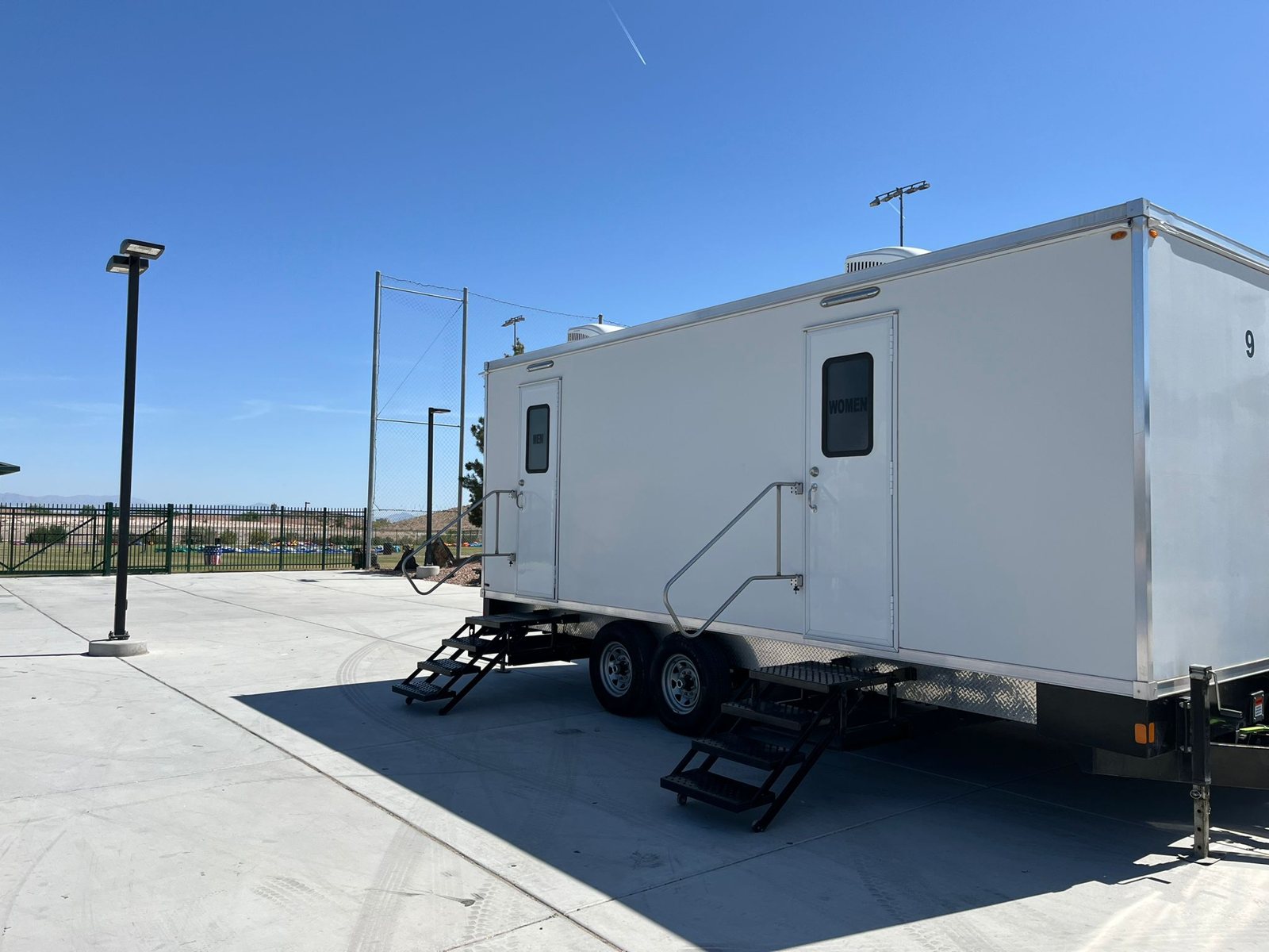 A Lavatory 9-Station restroom trailer with steel stairs on a concrete plaza beside ball fields, a tall backstop net and light poles