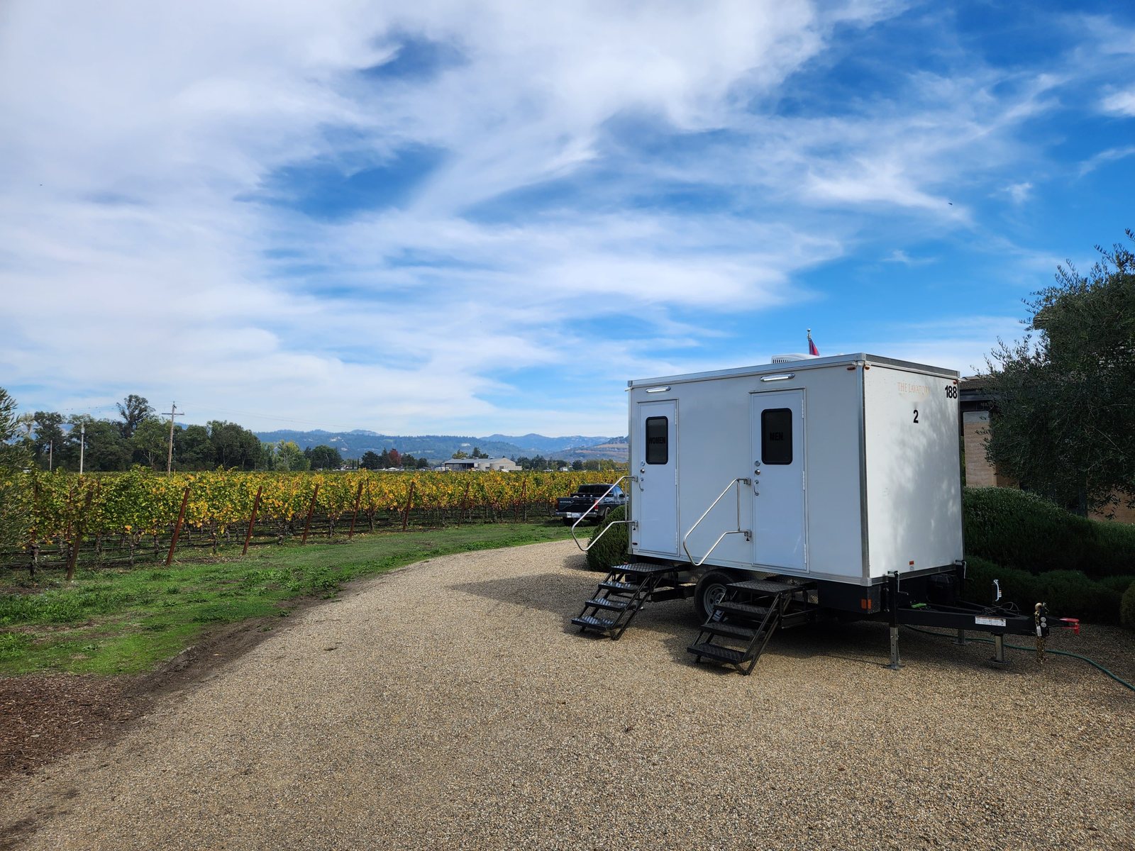 A Lavatory 2-Station luxury restroom trailer with separate women's and men's doors and steel stairs on a gravel drive beside a vineyard, with hills in the distance