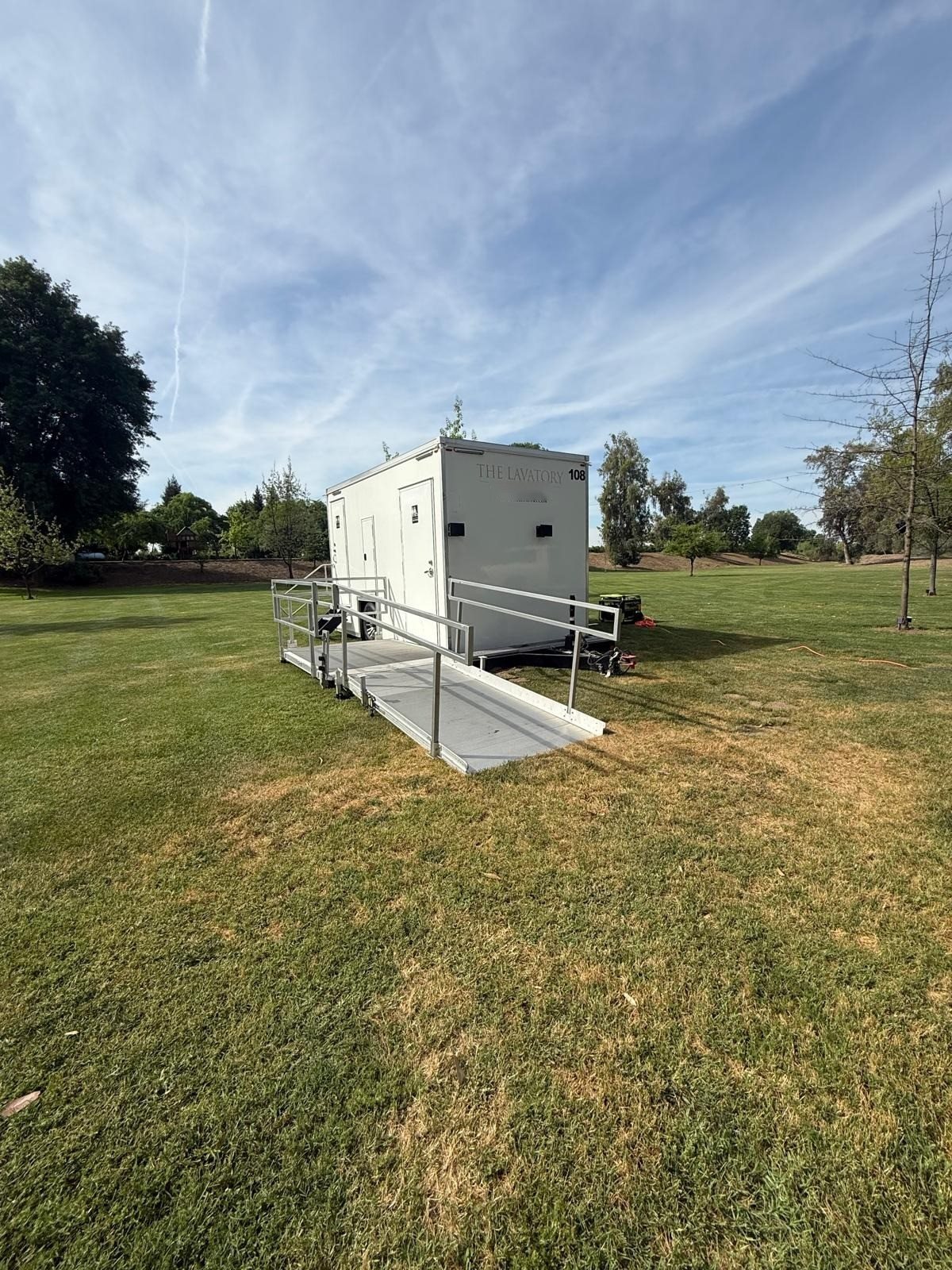 A Lavatory ADA restroom trailer with an aluminum ramp and handrails set on a wide grass lawn