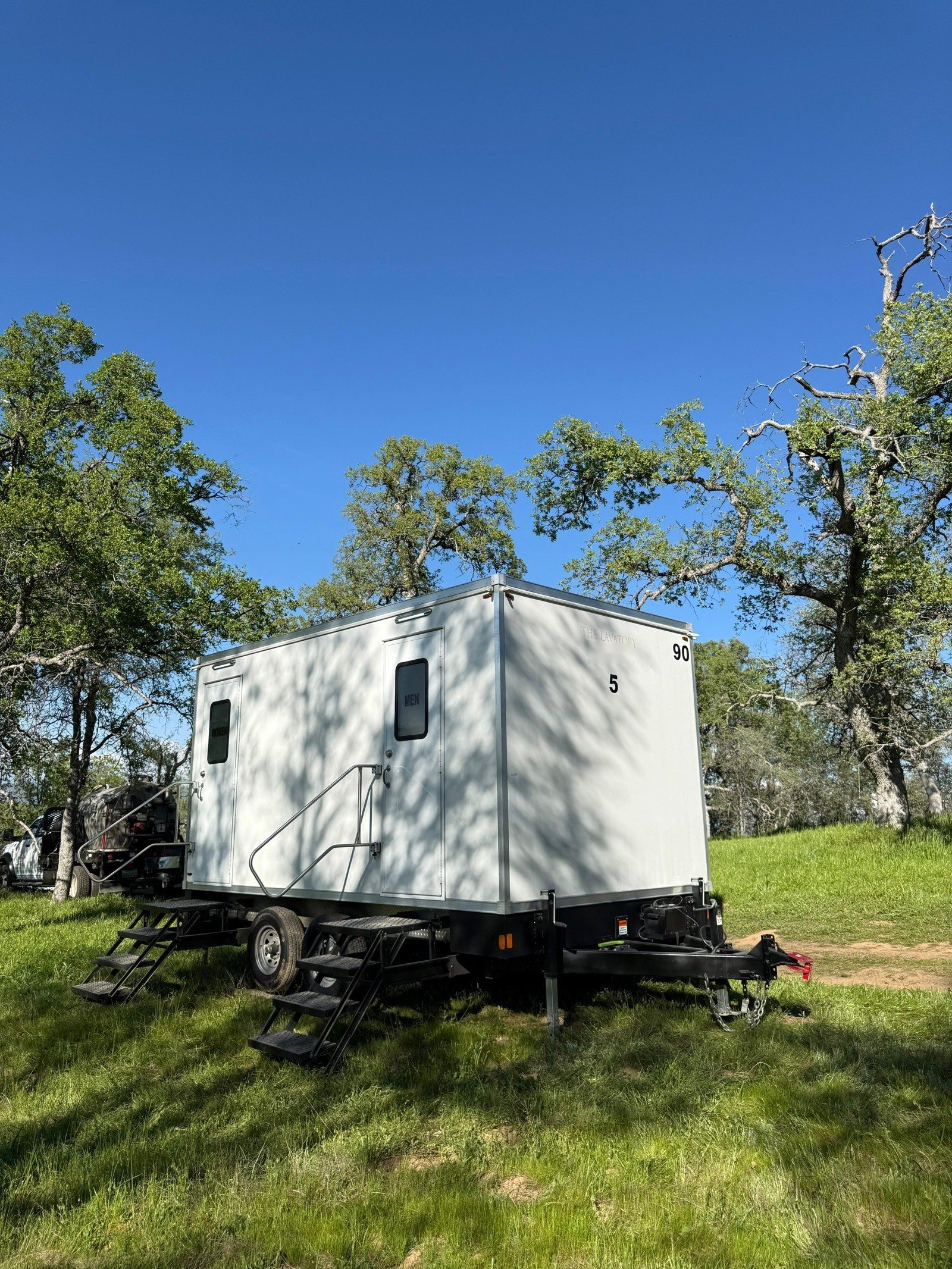 A Lavatory 5-Station restroom trailer parked on green grass under big oak trees on open ranch land