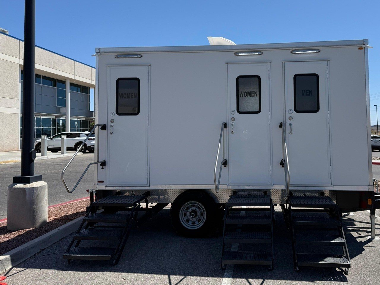 A Lavatory 3-Station restroom trailer with two women's doors and one men's door in an office building parking lot