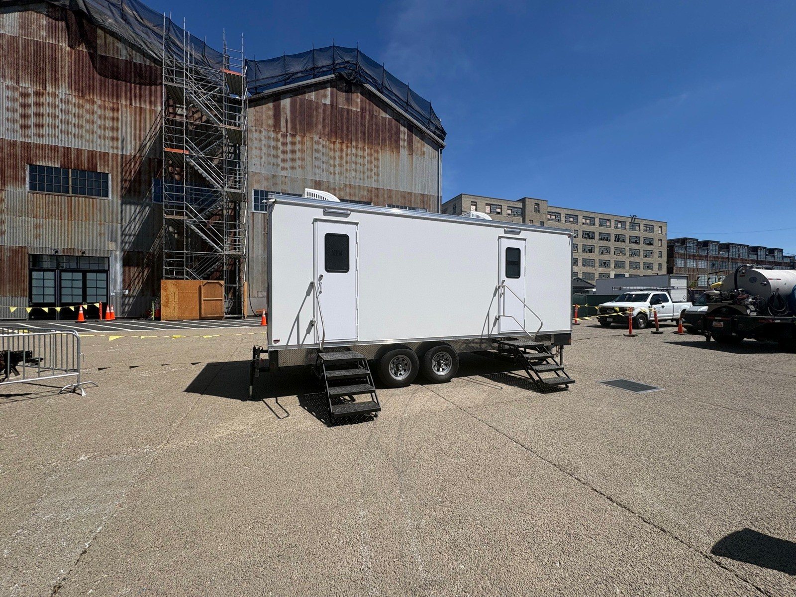 A Lavatory restroom trailer with steel stairs on a paved lot beside an old brick and metal warehouse wrapped in scaffolding