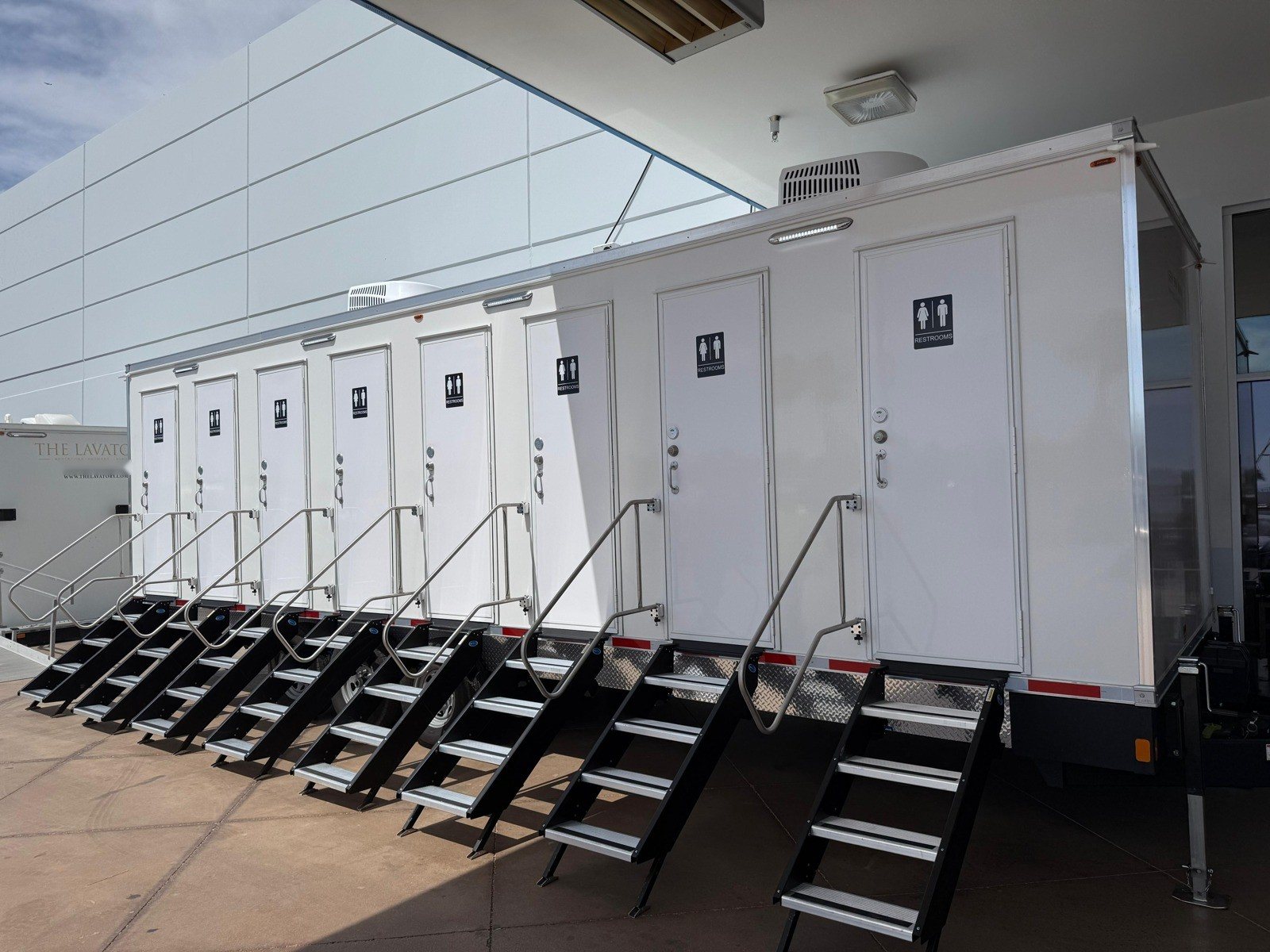 A Lavatory 8-Station private stall restroom trailer with eight lockable doors and steel stairs parked under a building overhang