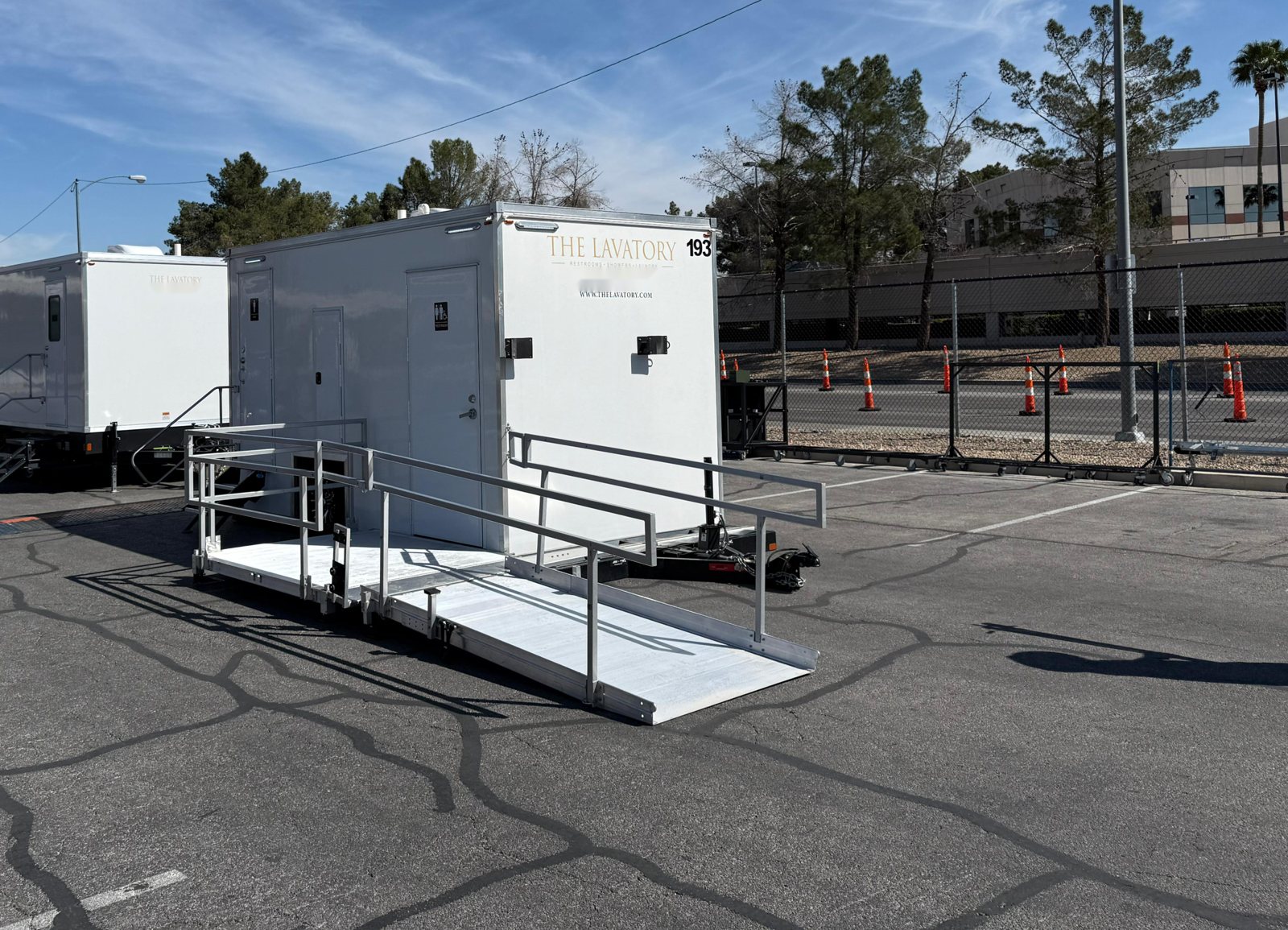 A Lavatory ADA restroom trailer with an aluminum ramp and handrails set up in a paved parking lot next to a second restroom trailer