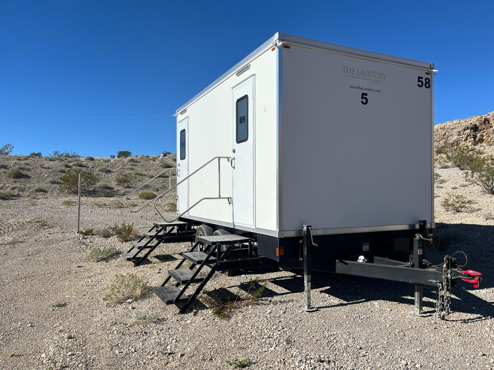 A Lavatory 5-Station restroom trailer on rocky desert ground with creosote bushes and a rock hillside behind it