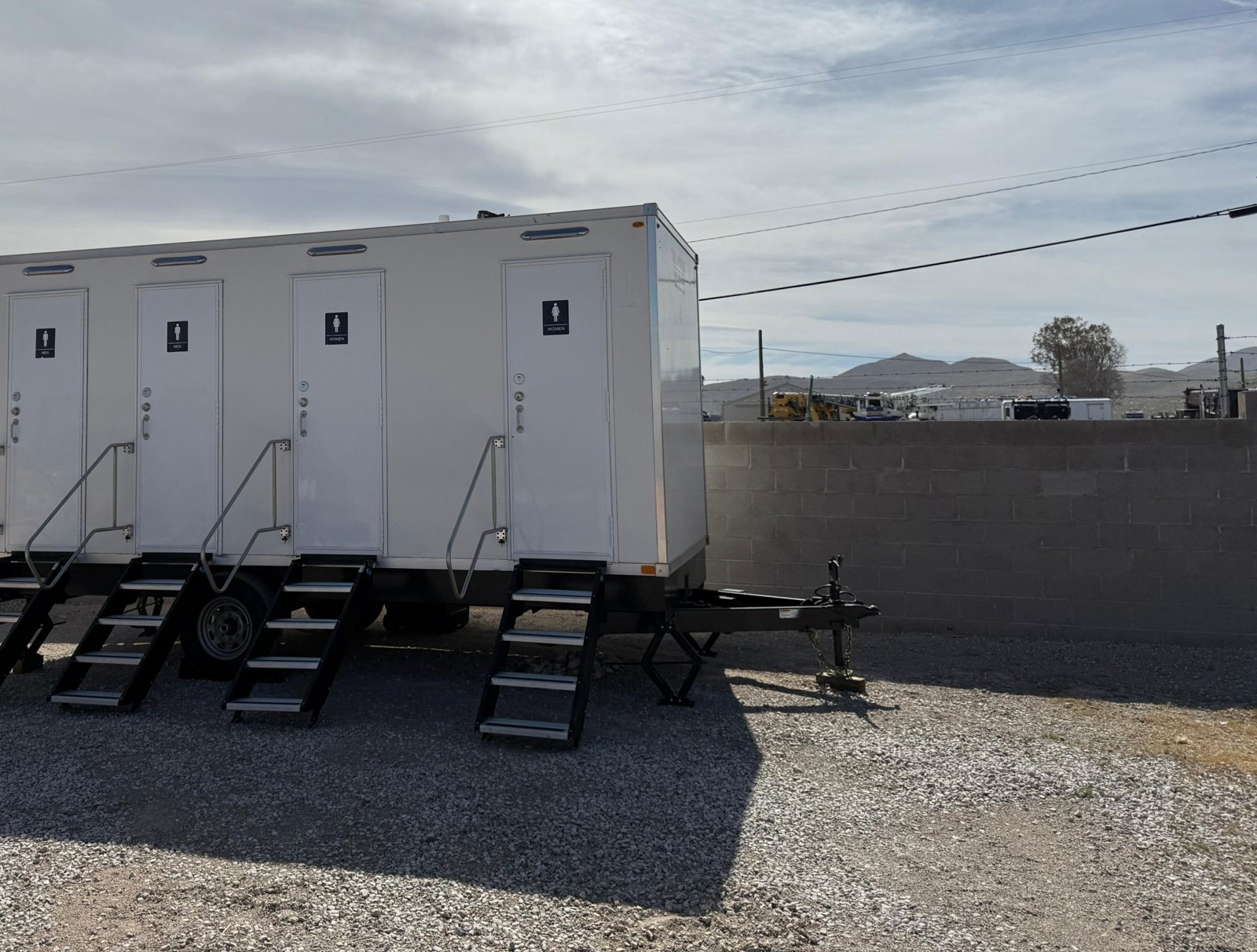 A Lavatory restroom trailer with four doors and four sets of steel stairs on a gravel lot in front of a block wall