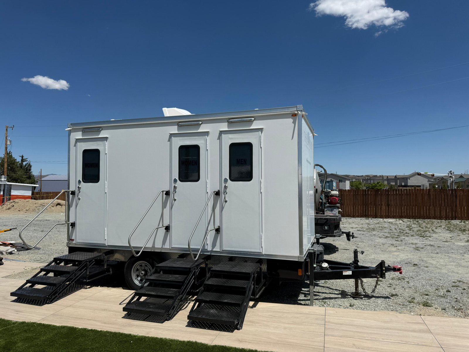 A Lavatory 3-Station restroom trailer with steel stairs on a gravel lot beside a sidewalk, with new homes and a wood fence behind it