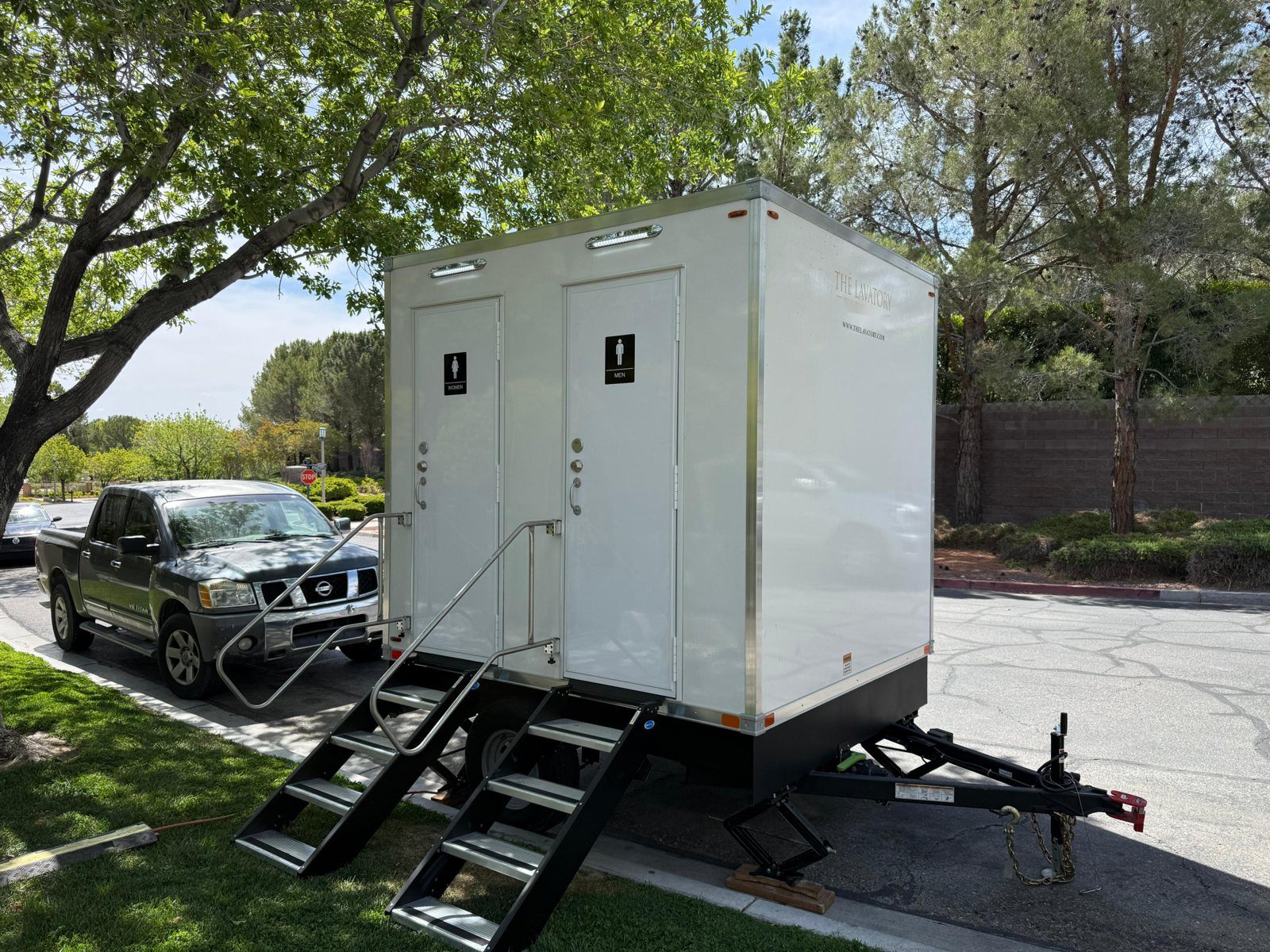 A Lavatory 2-Station restroom trailer with women's and men's doors parked at a curb under shade trees beside a lawn