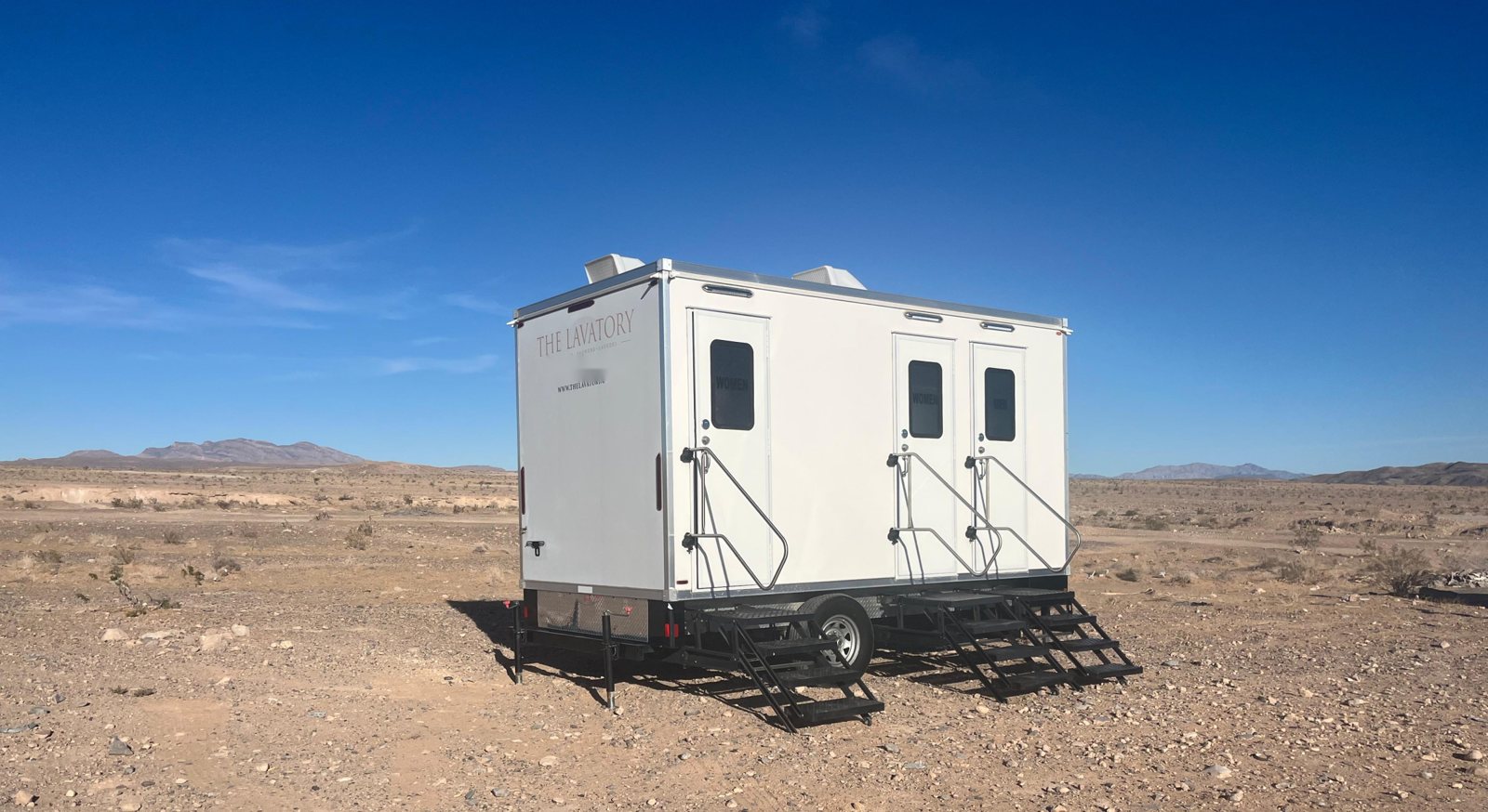 A Lavatory 3-Station restroom trailer with two women's doors, one men's door and steel stairs set on open desert ground with mountains on the horizon