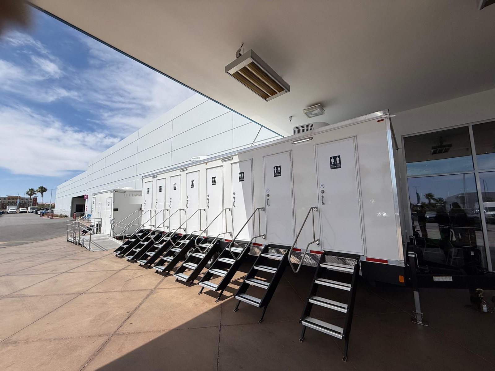 A row of Lavatory private stall restroom trailers with steel stairs and an ADA restroom trailer with a ramp beside a large event building