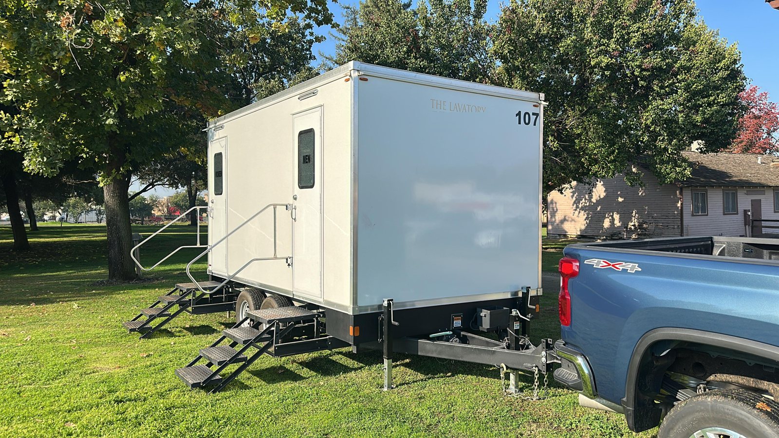 A Lavatory restroom trailer with steel stairs and handrails parked on a green lawn under big shade trees