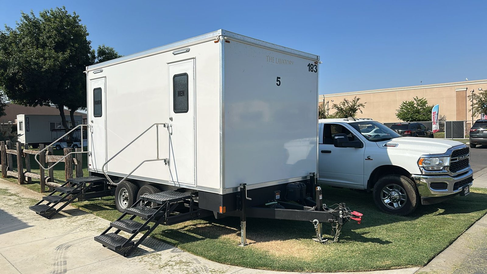 A Lavatory 5-Station restroom trailer on a grass strip beside a white work truck at a commercial lot