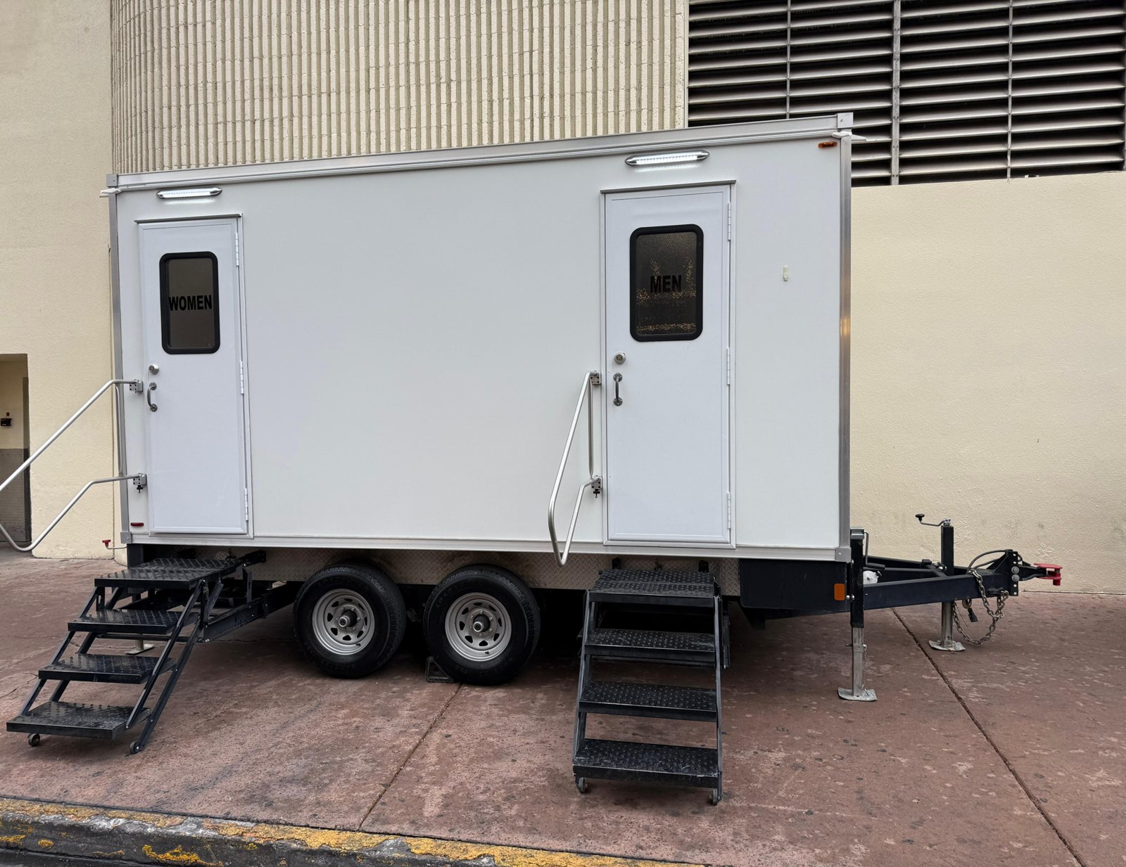 A Lavatory luxury restroom trailer with separate women's and men's doors, two steel stair sets and handrails, parked on a sidewalk beside a building